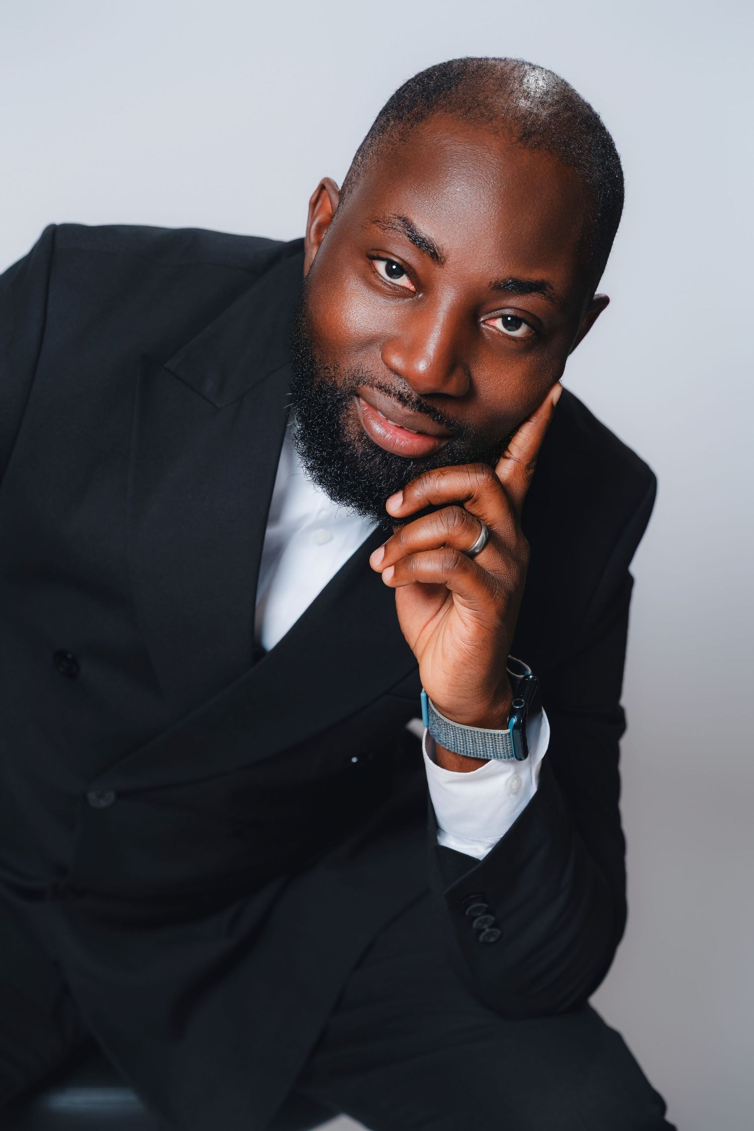 Professional portrait of someone in a black suit and tie with hand thoughtfully placed near their face against gray background.