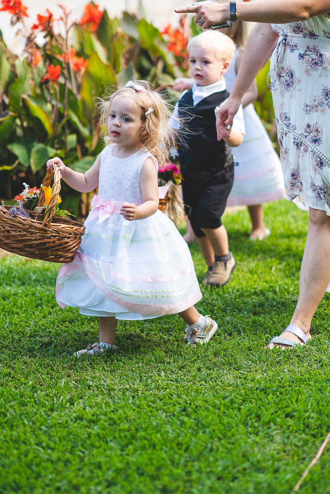 Young flower girls in white dresses walking on green grass during an outdoor wedding ceremony.