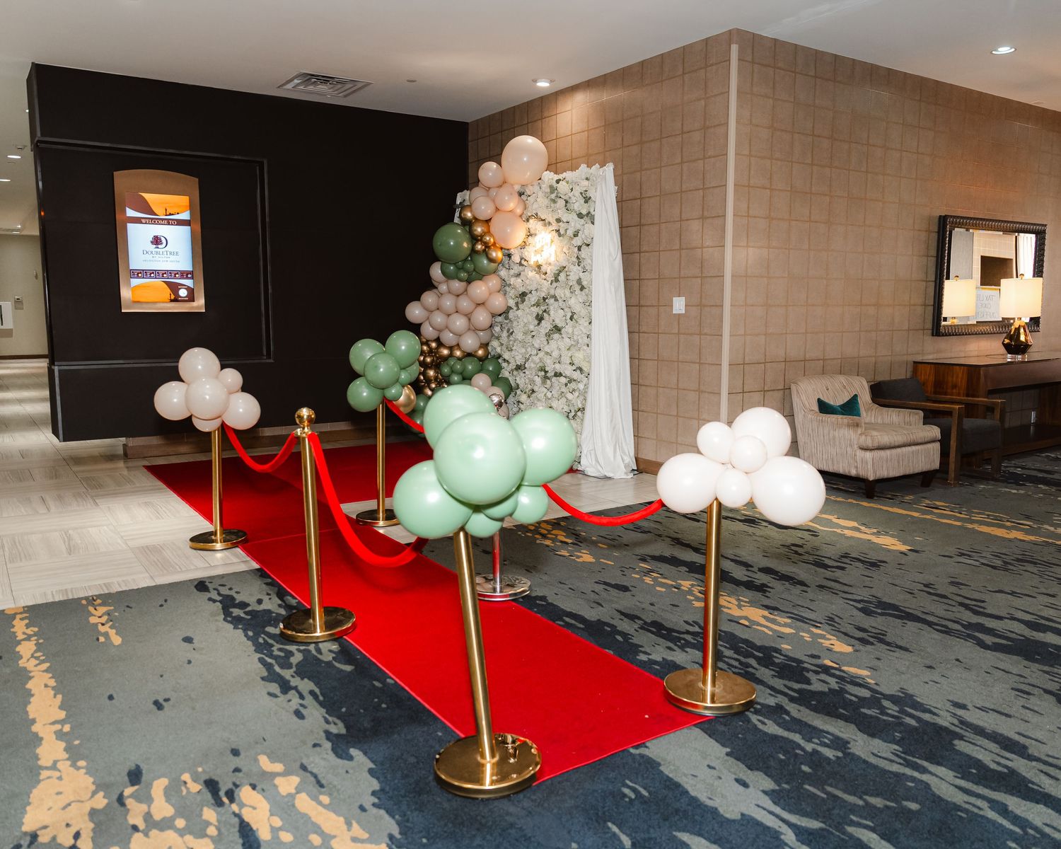 Elegant event setup with red carpet, balloon decorations in mint green and peach colors, and gold stanchions in a hotel lobby.