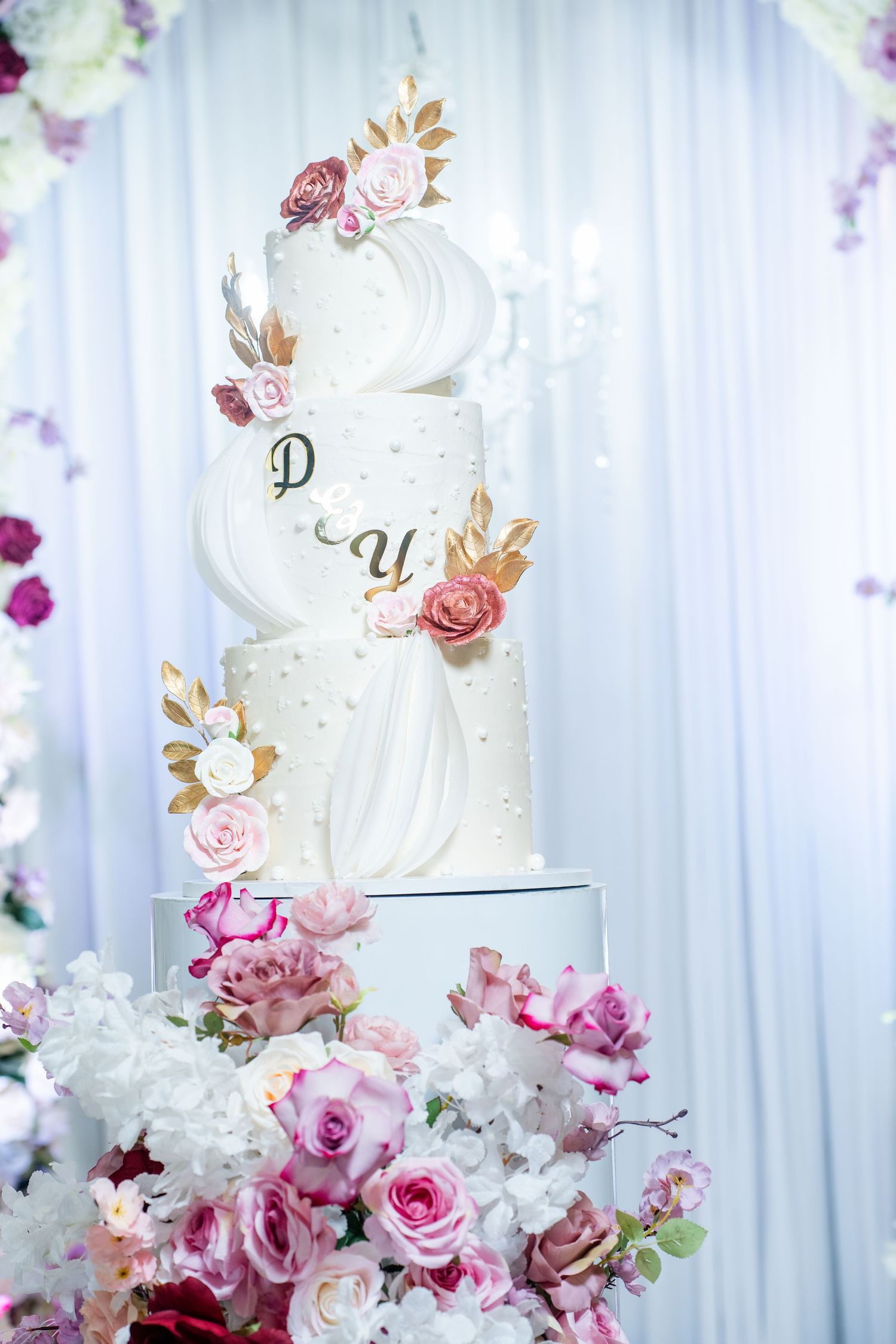 Elegant white wedding cake decorated with pink and purple flowers against a white draped backdrop.