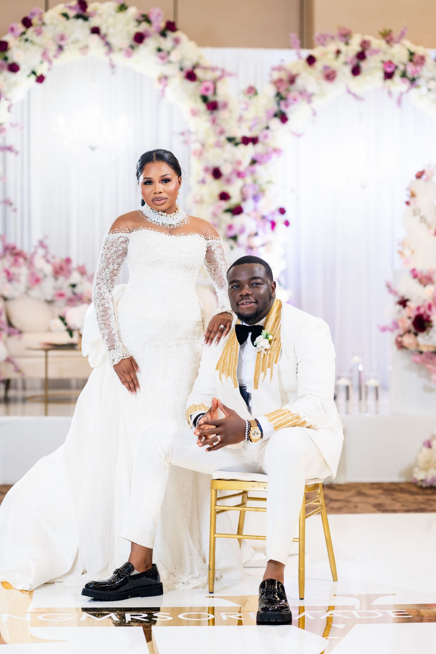 Newlyweds pose together on gold chairs with romantic floral wall backdrop in wedding attire.