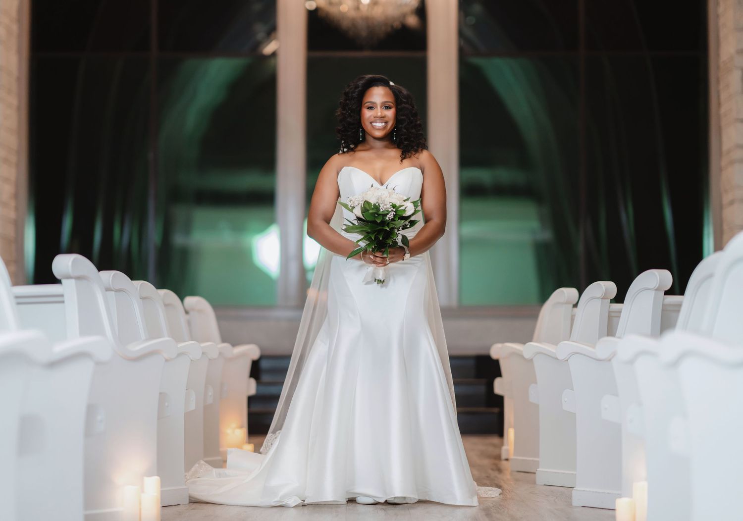 Bride poses elegantly in white gown inside venue with white chairs and dramatic lighting.