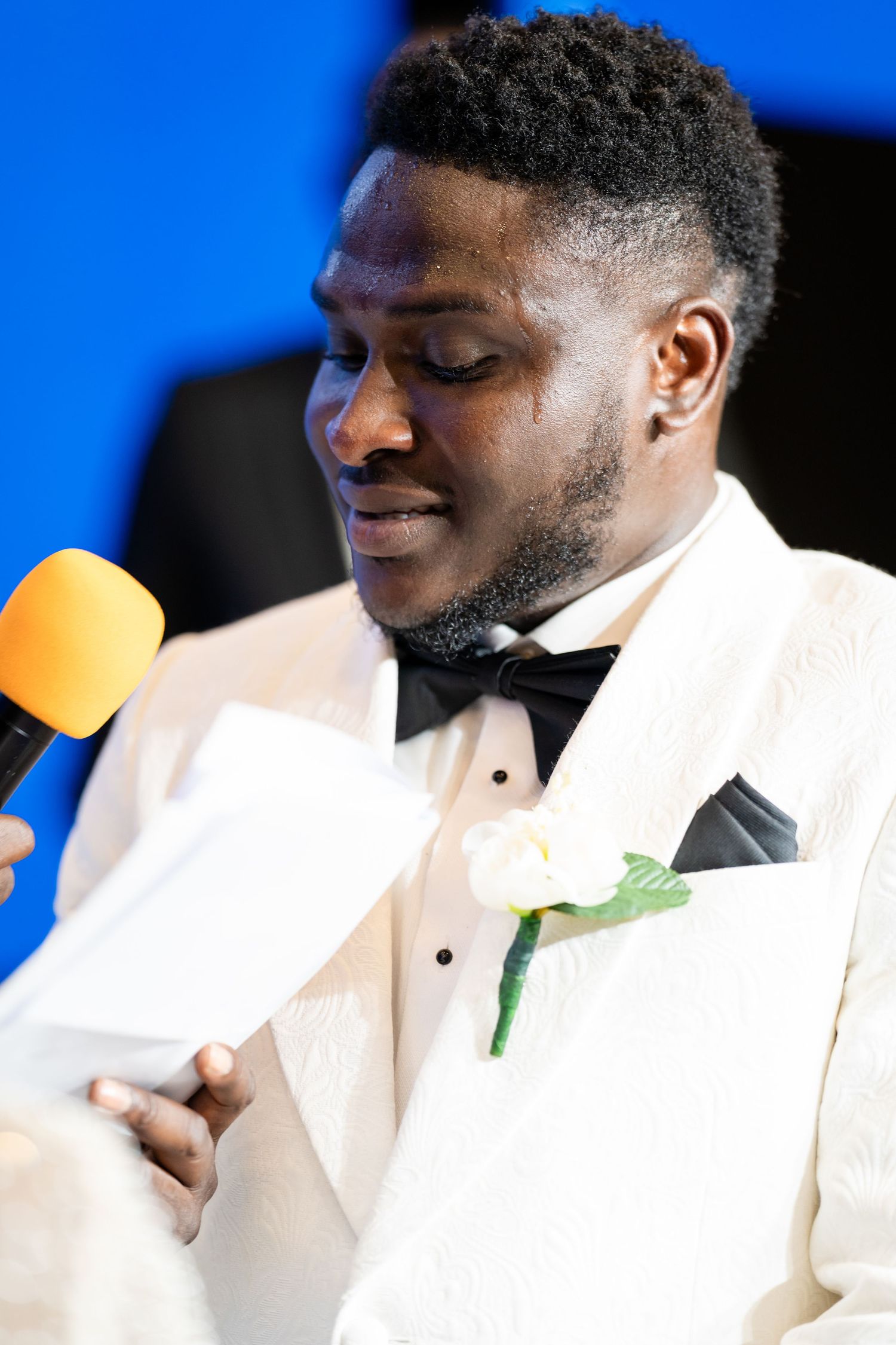 A groom in white tuxedo speaks into a yellow microphone during wedding reception.