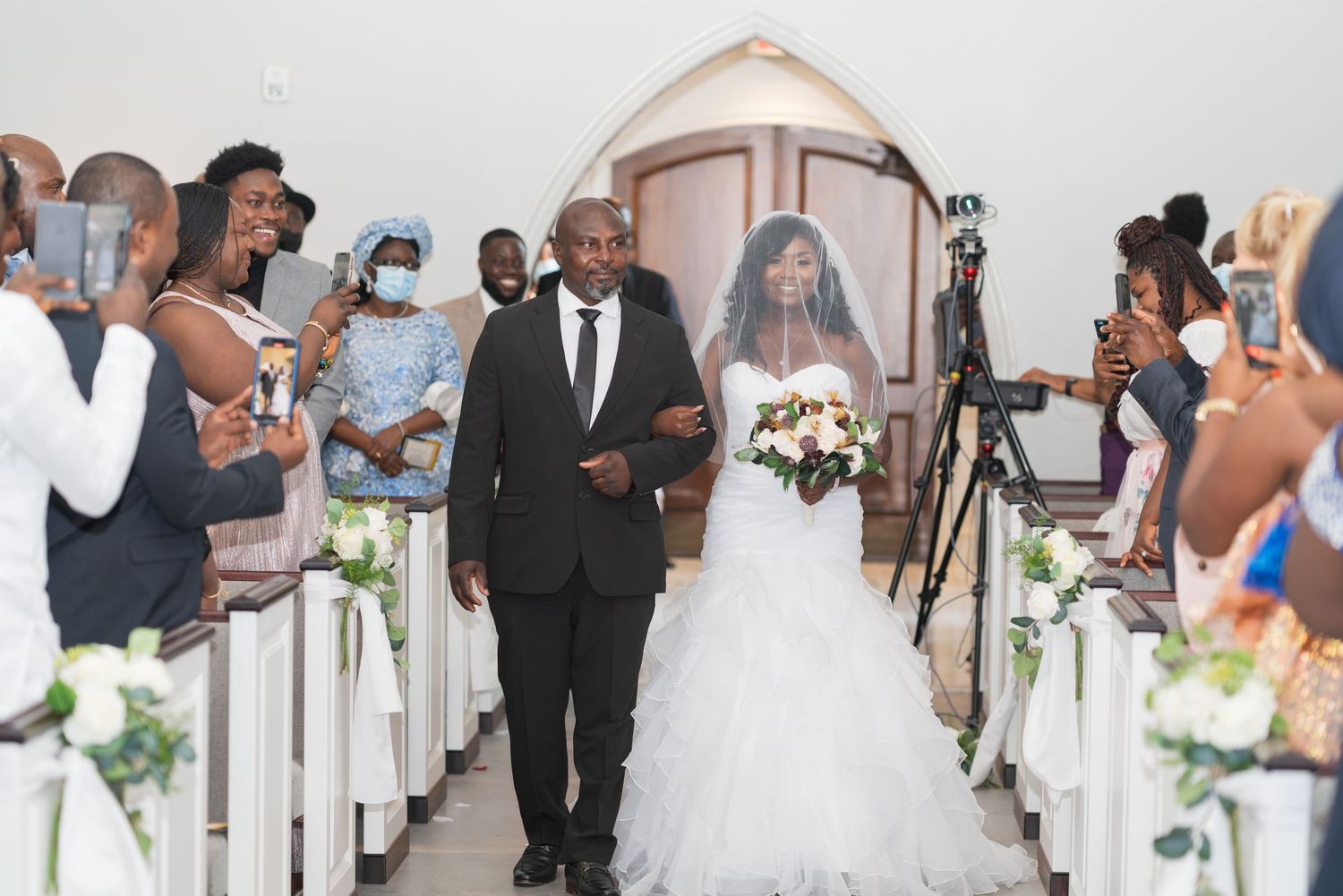 Wedding processional in a bright church with guests watching from decorated white pews.