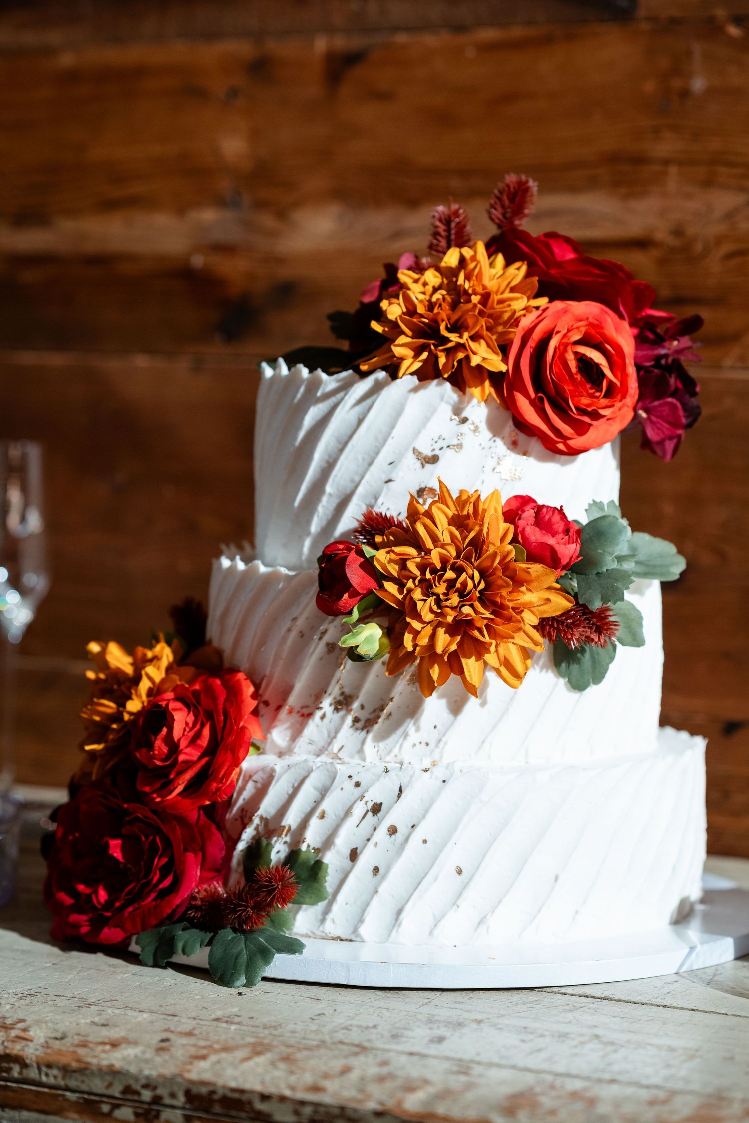 White tiered wedding cake decorated with burgundy and orange flowers on rustic wooden surface.