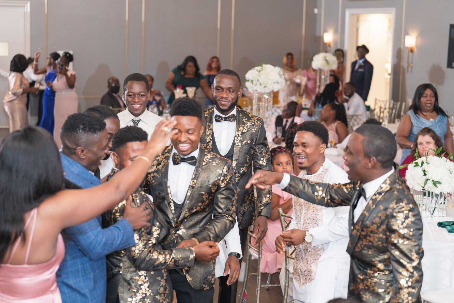 A celebratory moment at a wedding reception with groomsmen in gold patterned suits dancing and interacting with guests.