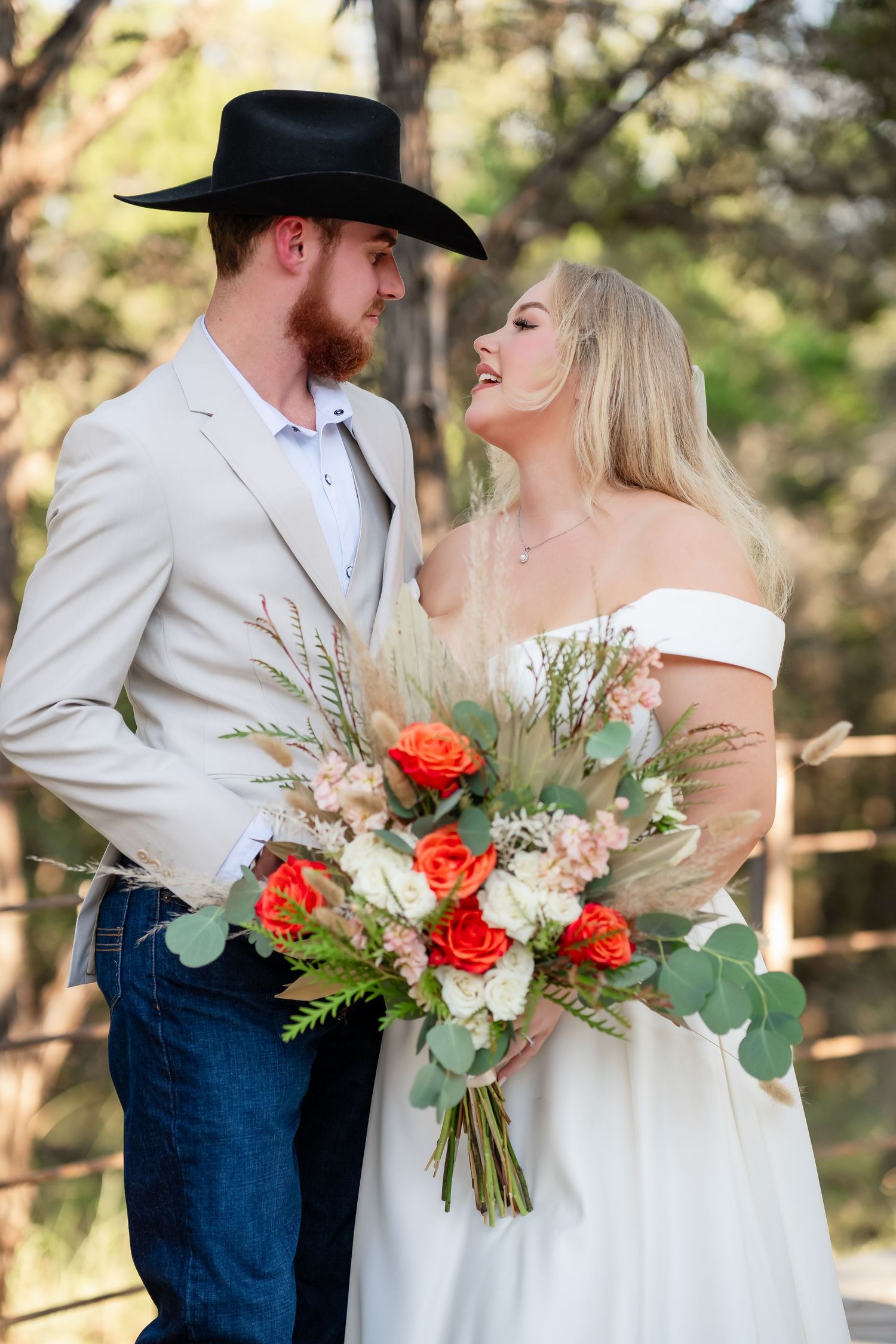 A romantic wedding photo with a couple in western attire holding a coral rose bouquet.
