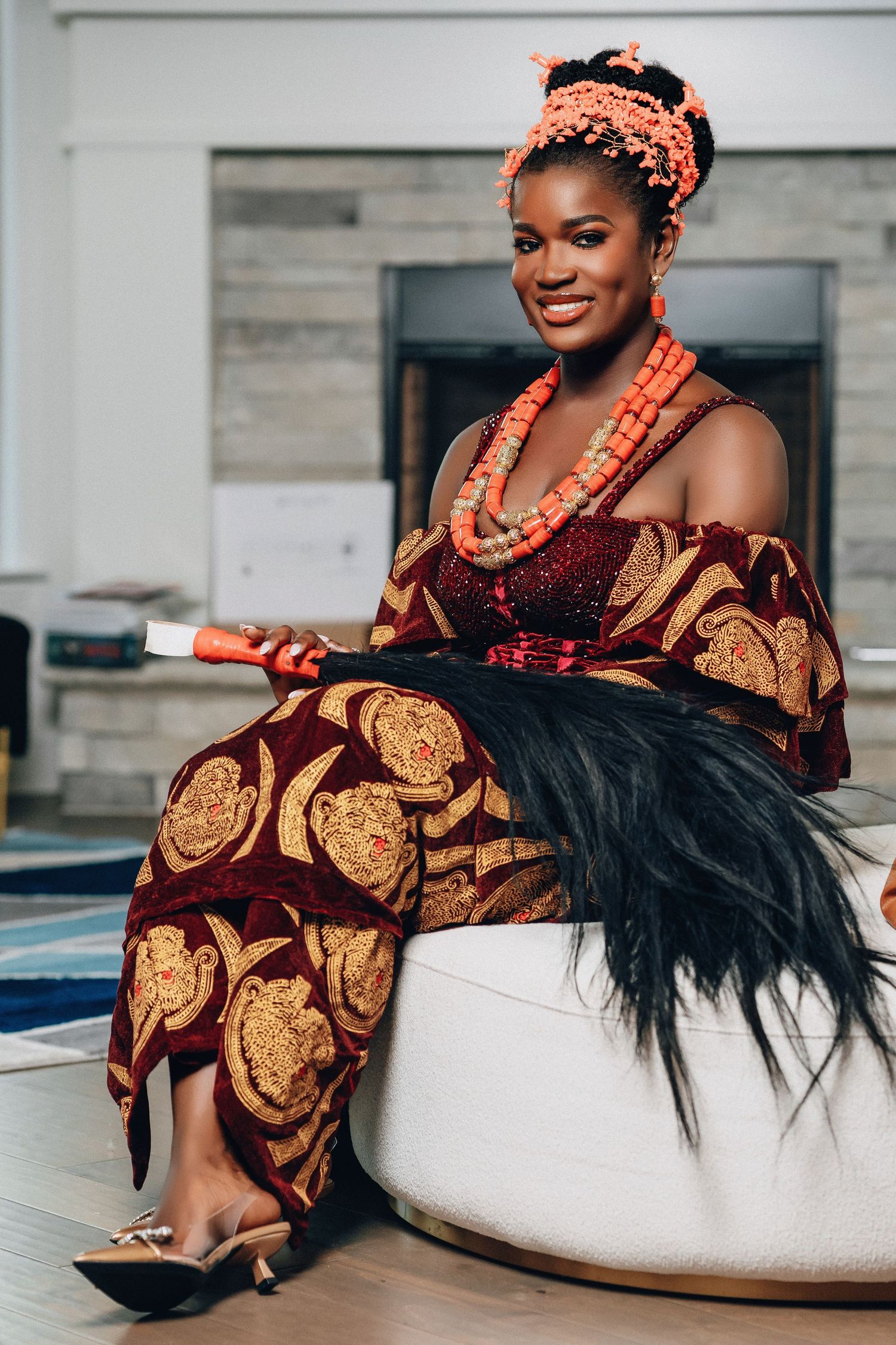 Traditional African fashion with coral beads and burgundy ankara print outfit on white ottoman.