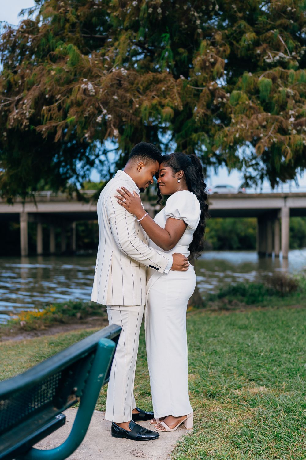 A romantic couple in white outfits embracing near a waterfront park with trees in the background.