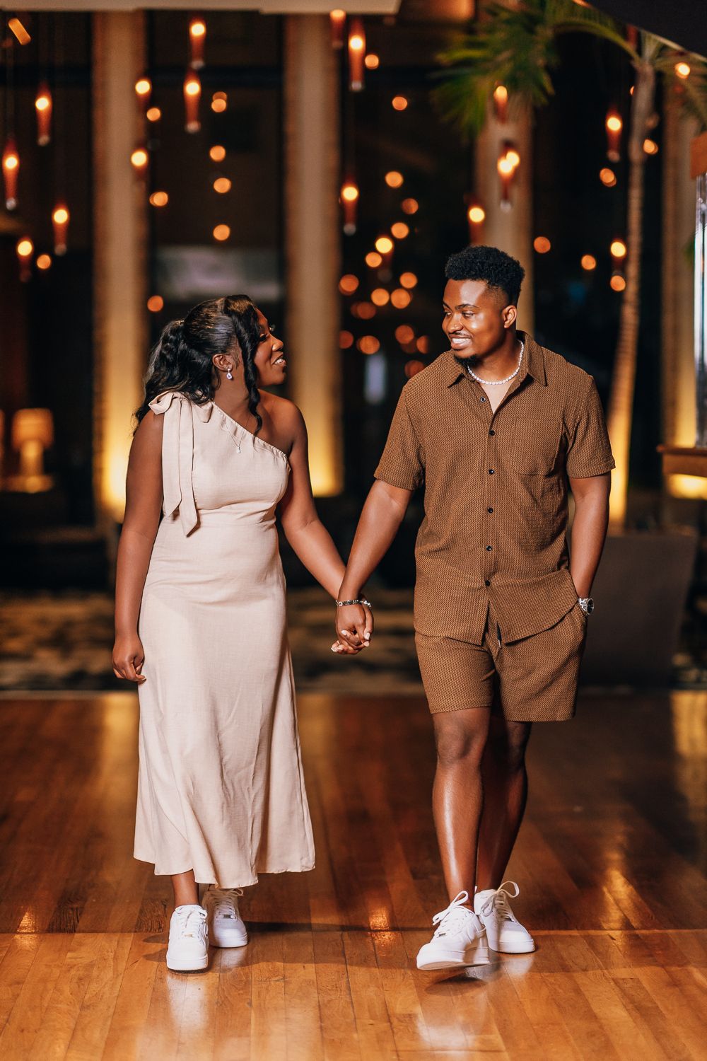 A couple in coordinated brown and beige outfits hold hands and smile while standing on a hardwood floor with twinkle lights.