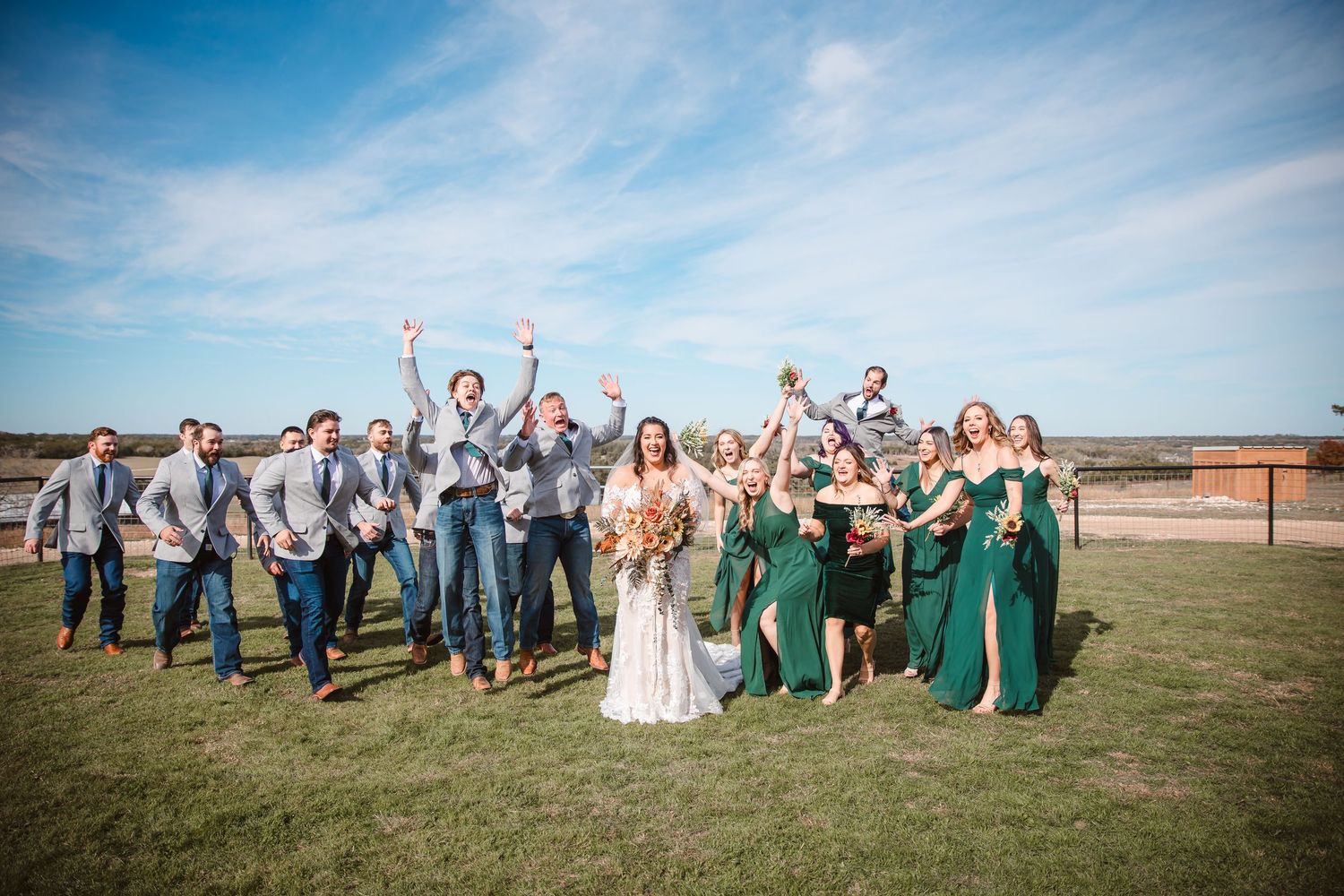Large wedding party poses together outdoors wearing green and gray attire against a bright blue sky.