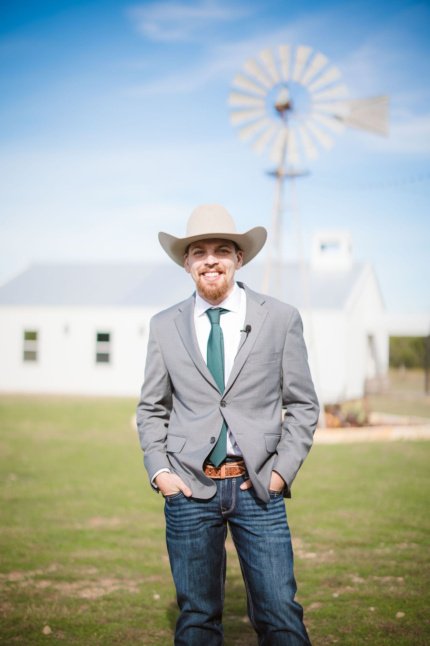 A western-style groom in a gray suit jacket, teal tie and cowboy hat poses against a bright blue sky with a windmill.
