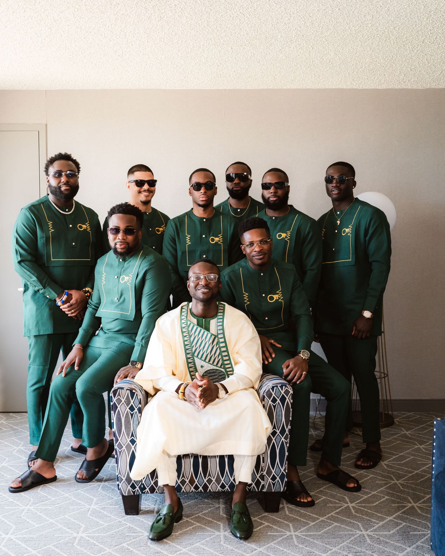 Wedding party wearing coordinated green suits posing together on patterned chairs.