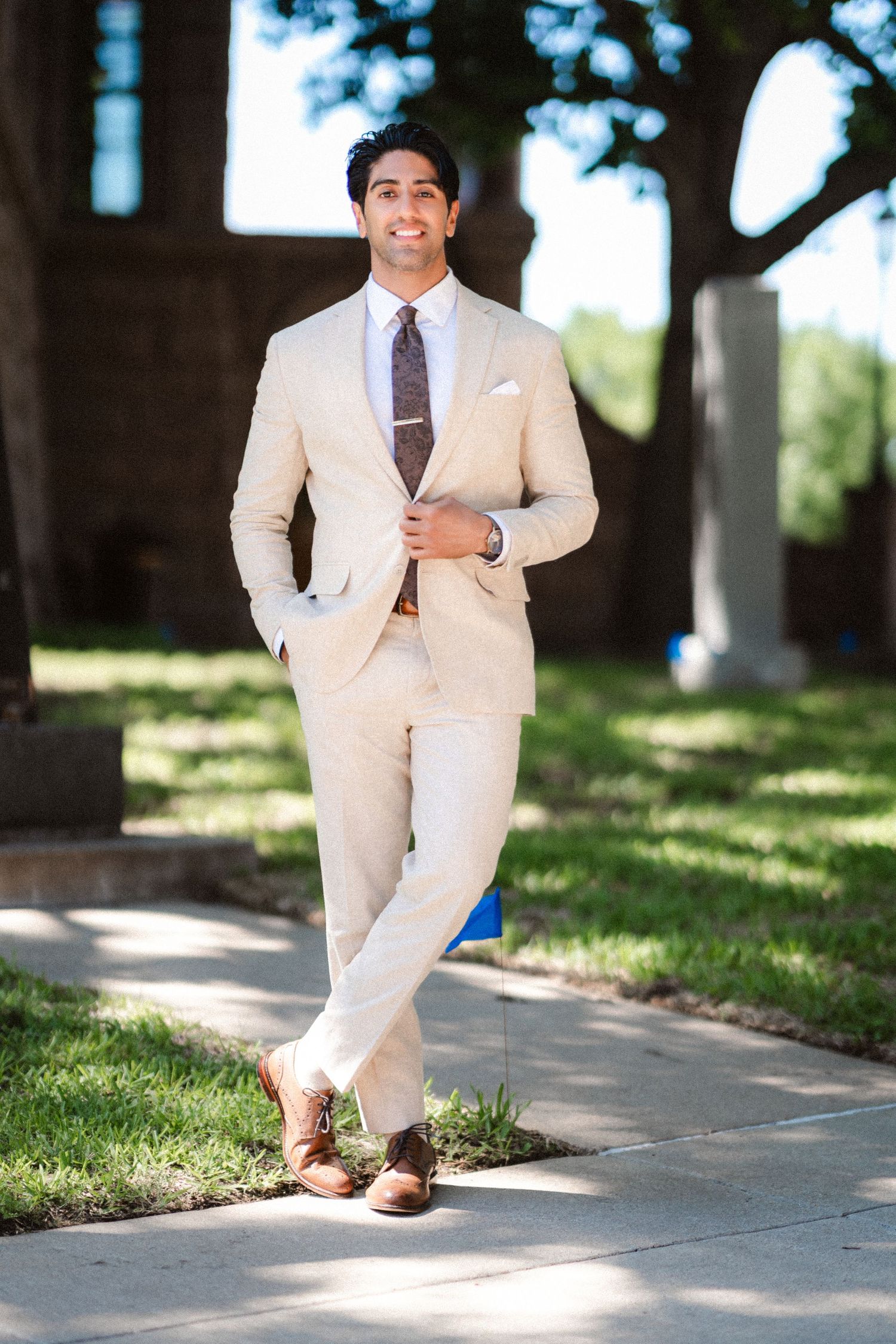 A groom in a cream colored suit and tie poses casually on a sunny pathway.