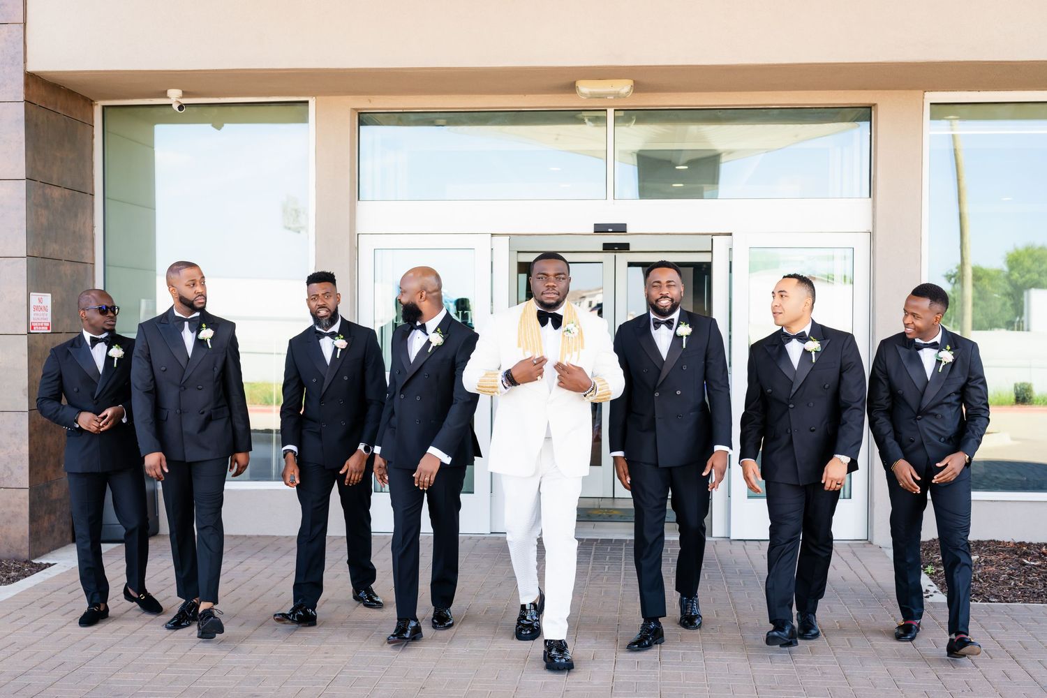 Wedding party members in black tuxedos and white dresses walk in line along a building exterior.