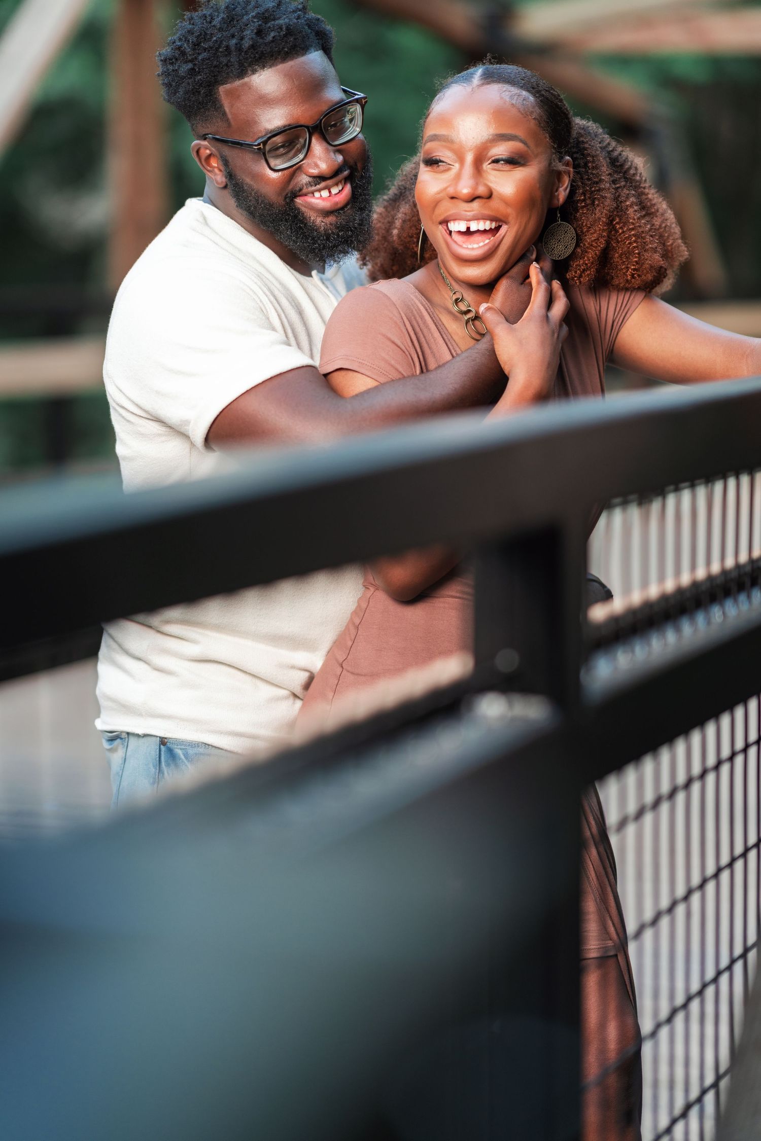 A couple shares joyful laughter while posing on a bridge railing.