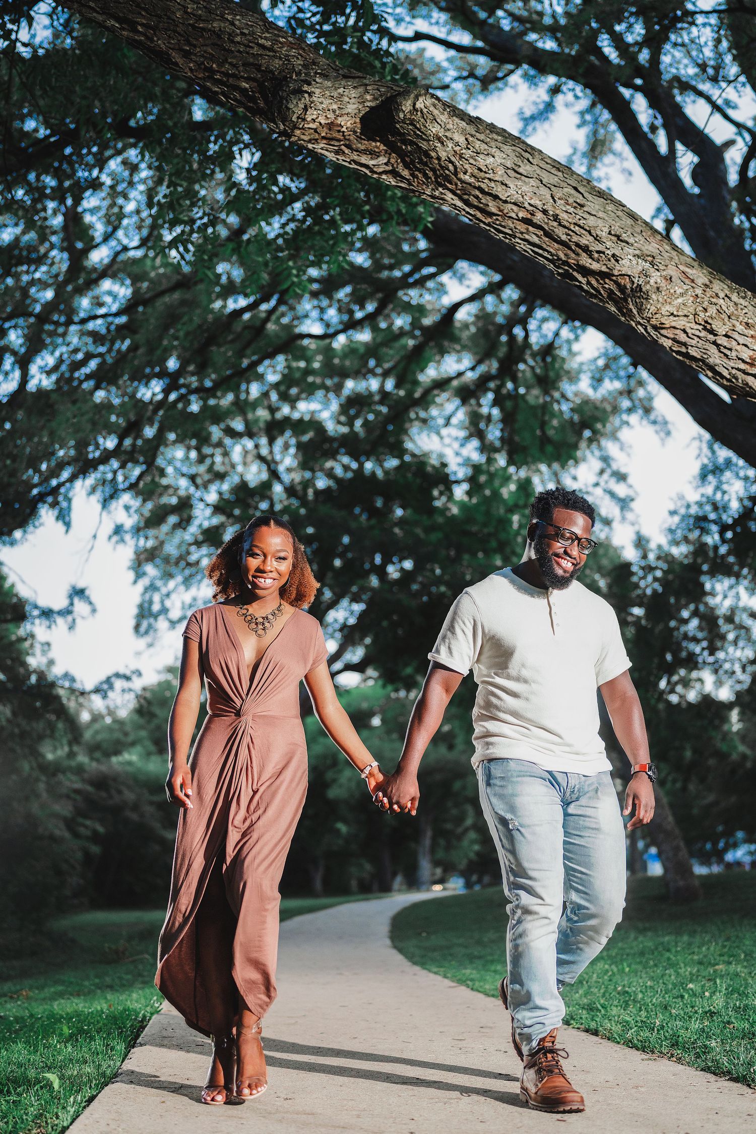 A couple walks hand in hand along a tree-lined path wearing casual outfits of brown dress and jeans on a sunny day.