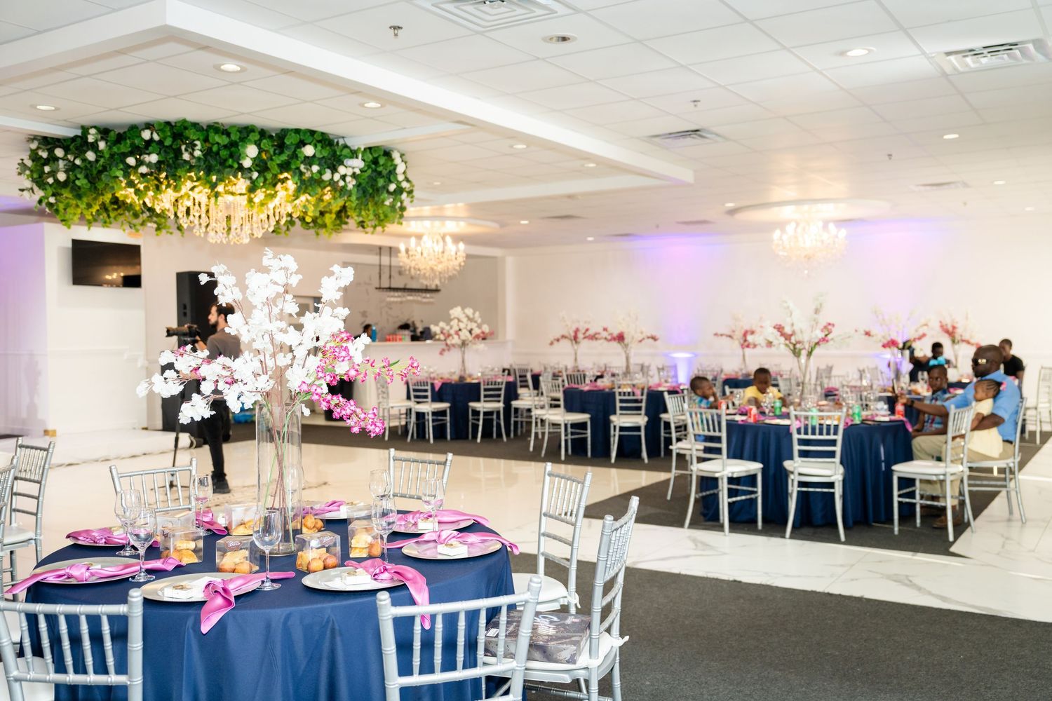 Modern white event venue decorated with greenery and purple lighting features elegant table settings and chiavari chairs.