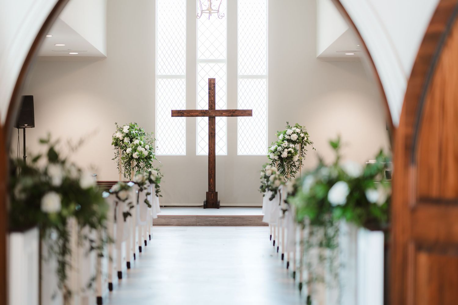 Bright church interior with wooden beams, white walls, and decorative greenery along the aisle.