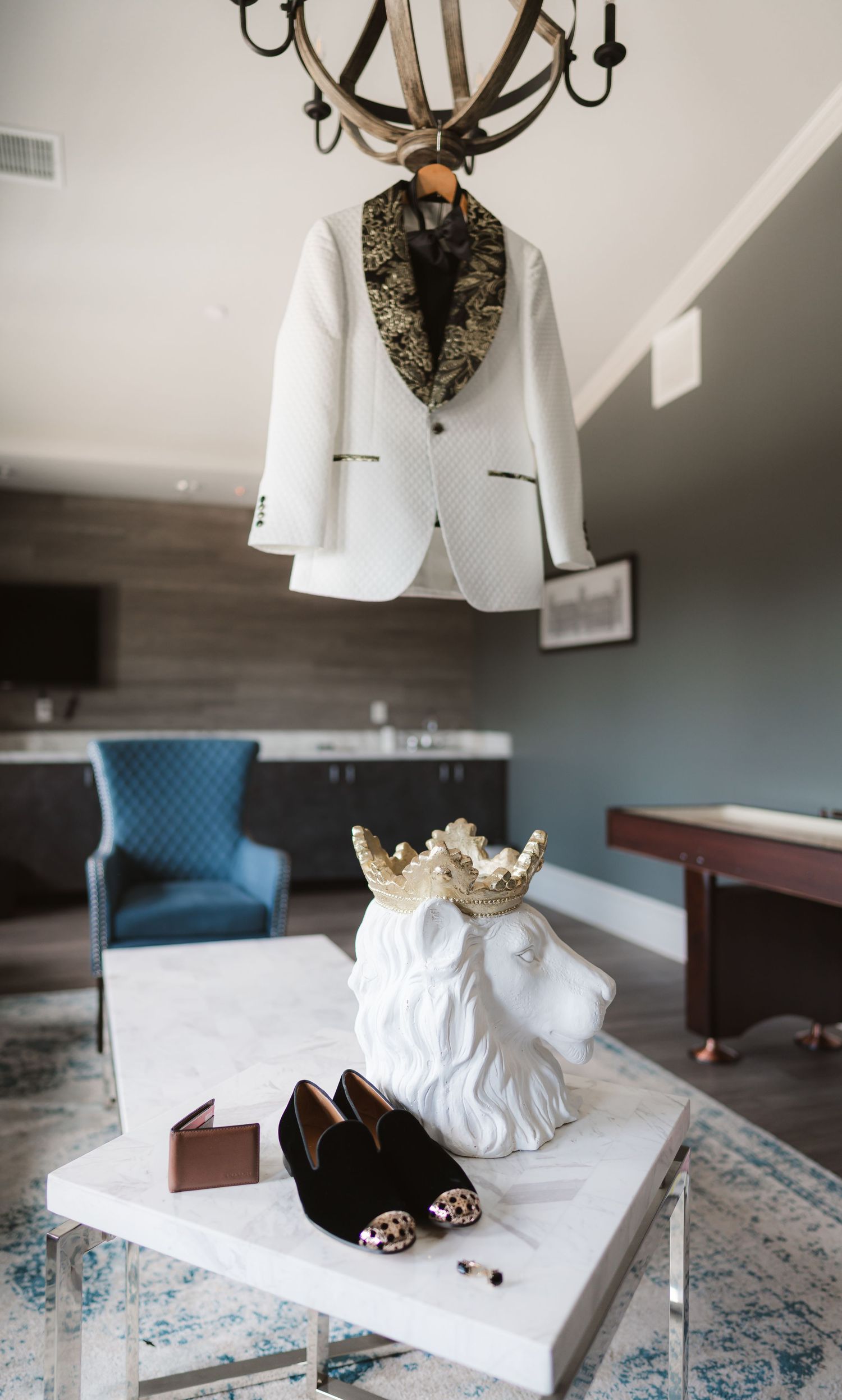 Wedding accessories including a white suit jacket, crown, and shoes arranged on a blue chair and carpet.