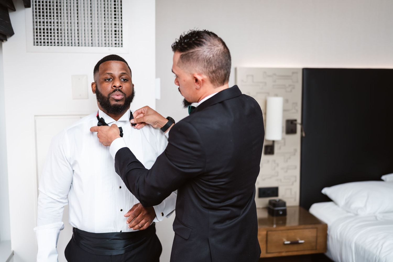 Wedding preparation scene showing formal attire adjustments in bright hotel room.