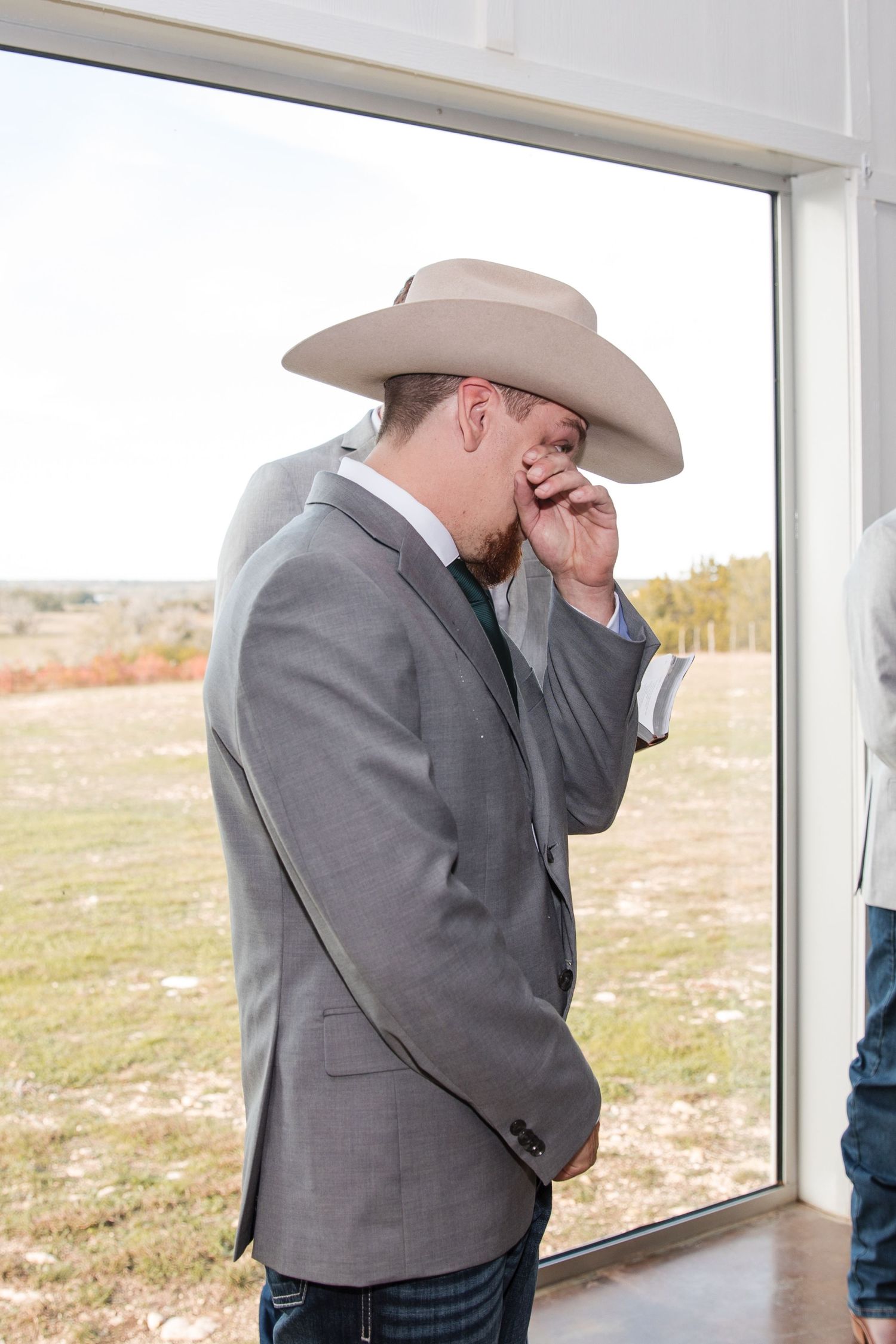 Emotional groom in a grey suit and cowboy hat wipes away tears during a first look moment.