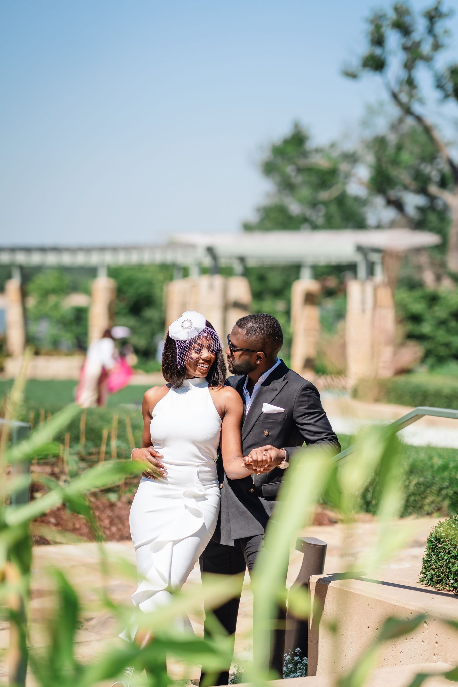 A couple shares an intimate moment in a lush garden setting with stone pillars and greenery.