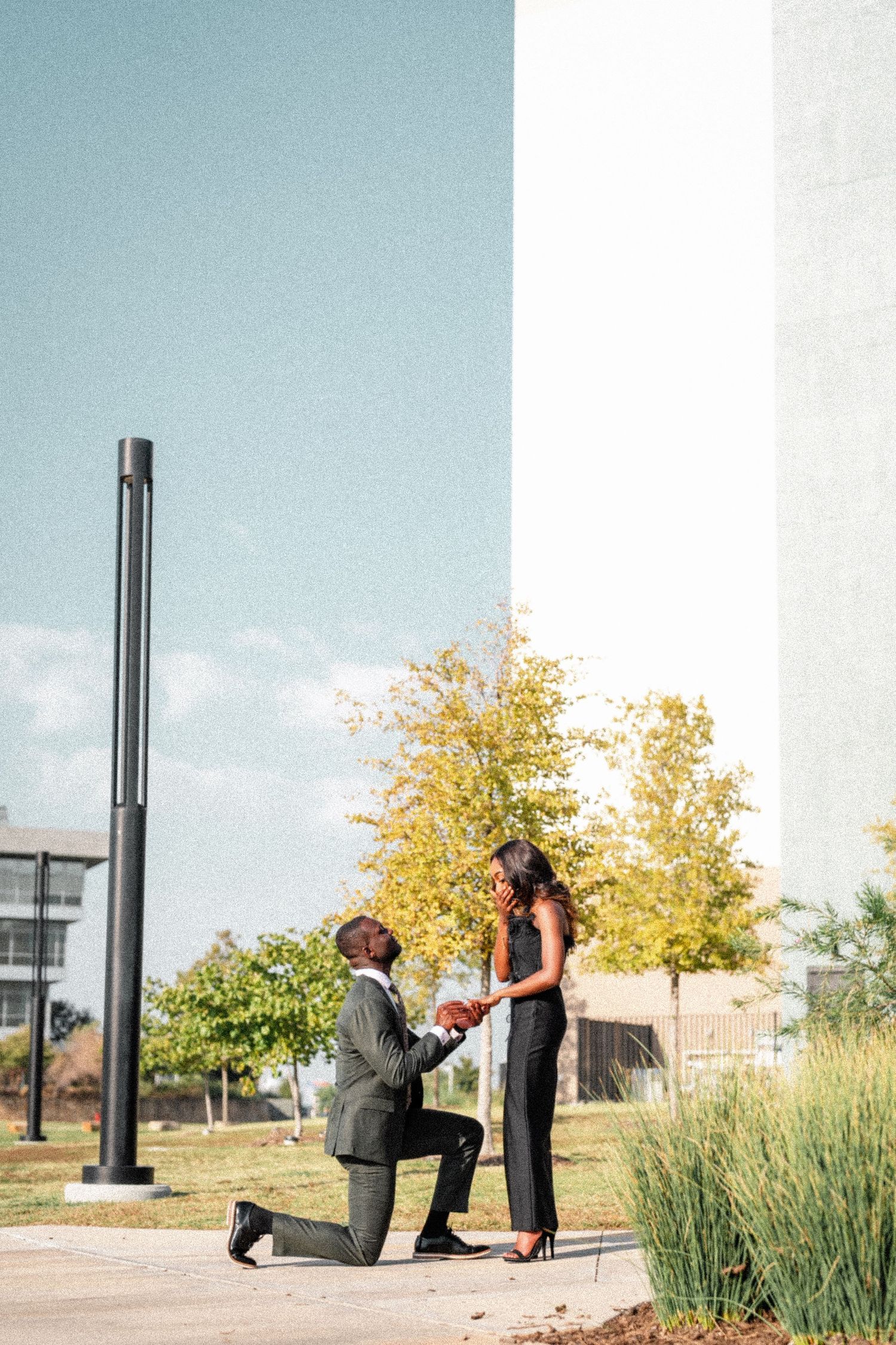 A romantic proposal scene unfolds on a walkway with autumn trees and blue sky in the background.
