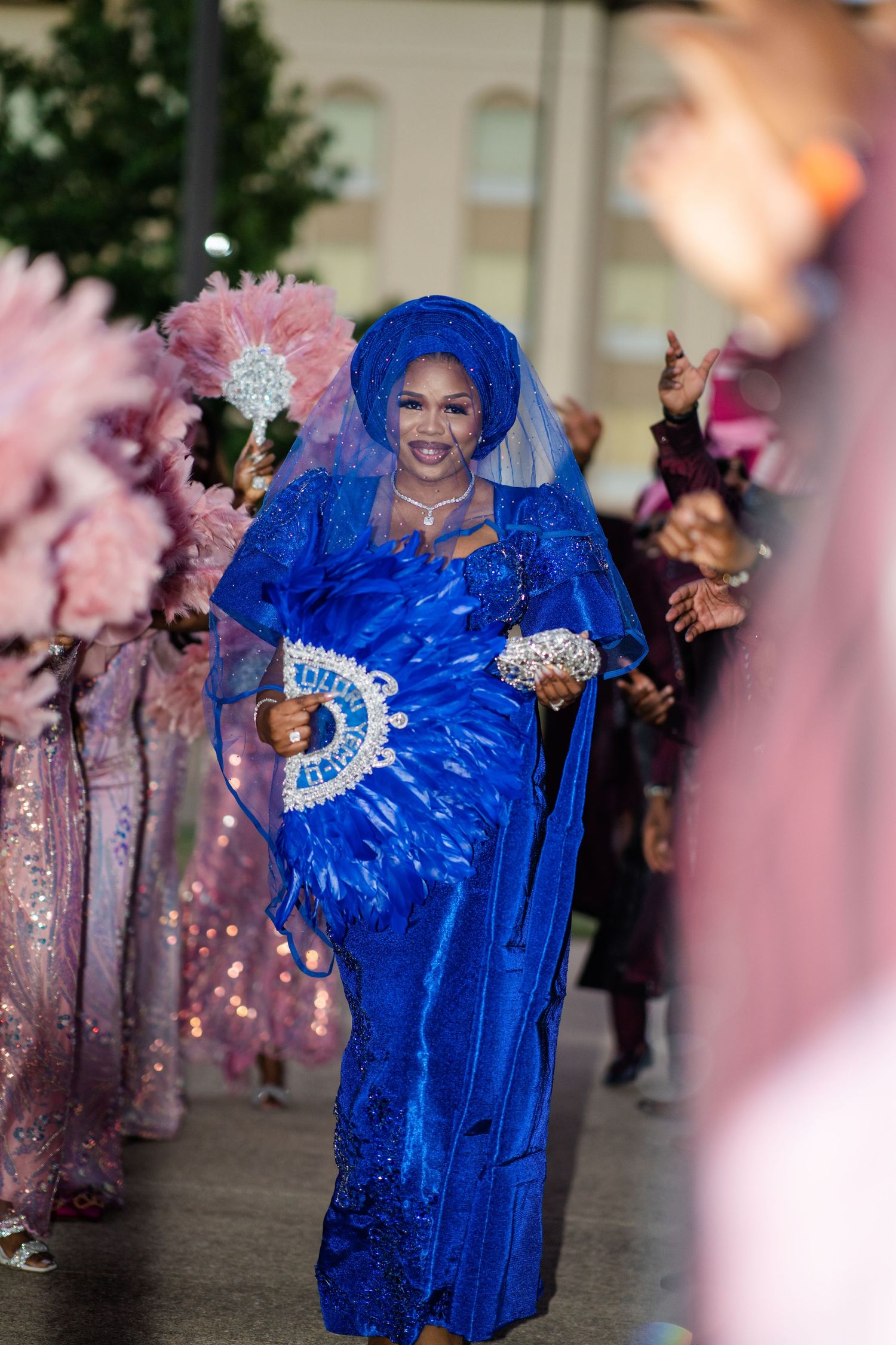 Wedding celebration with person in vibrant royal blue gown and feathers dancing joyfully among pink-dressed attendees.