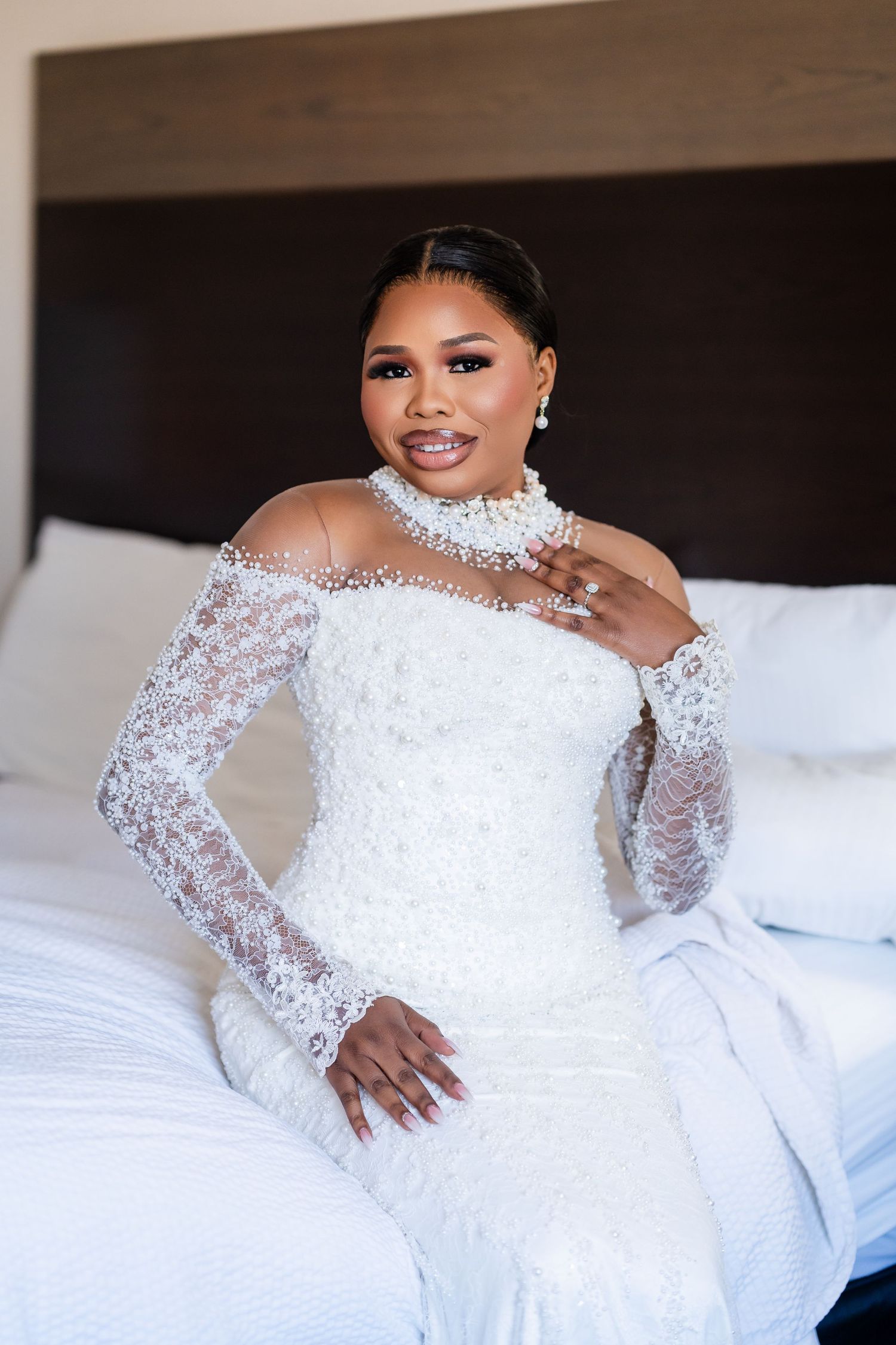Bride in intricate white lace wedding gown with crystal neckline poses on white bedding.