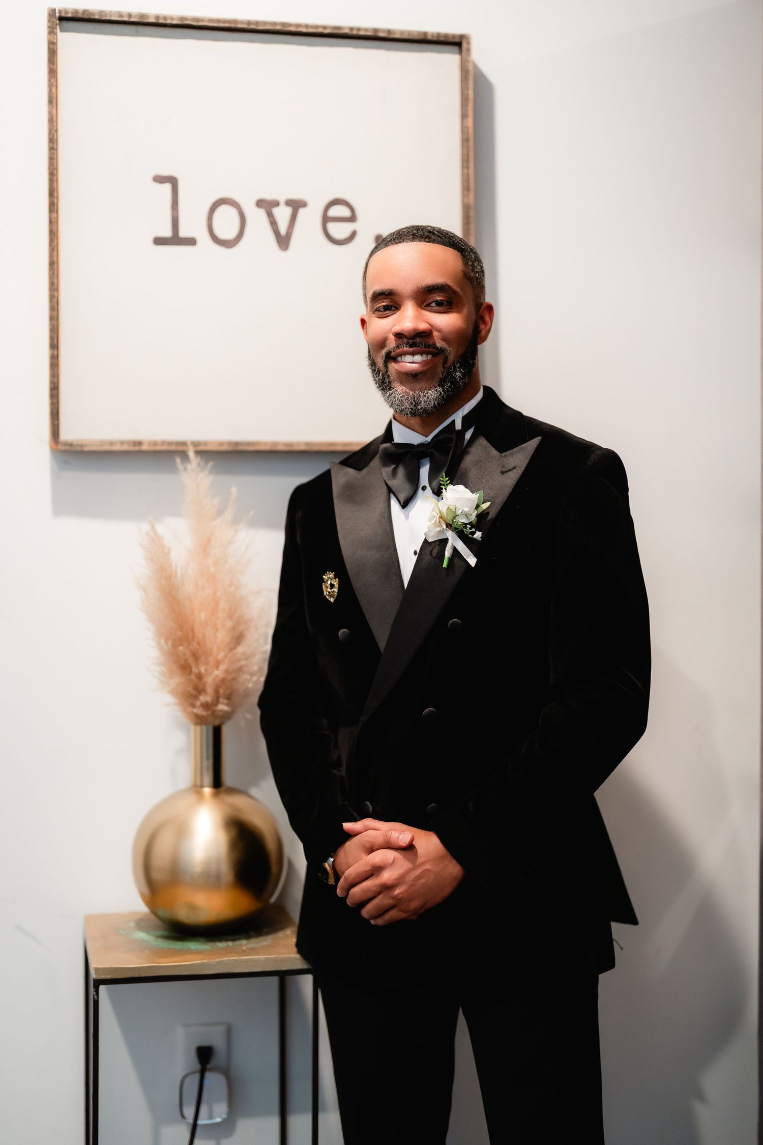 Two grooms in black tuxedos share a candid moment in front of a minimalist wall with 'love' decor.