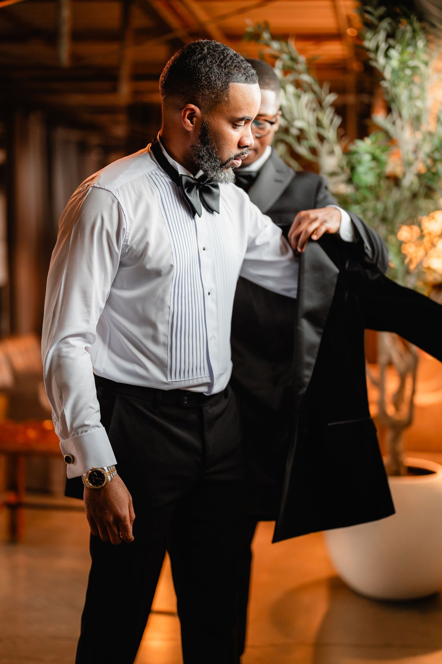 Groom adjusts formal tuxedo jacket in warm ambient lighting.
