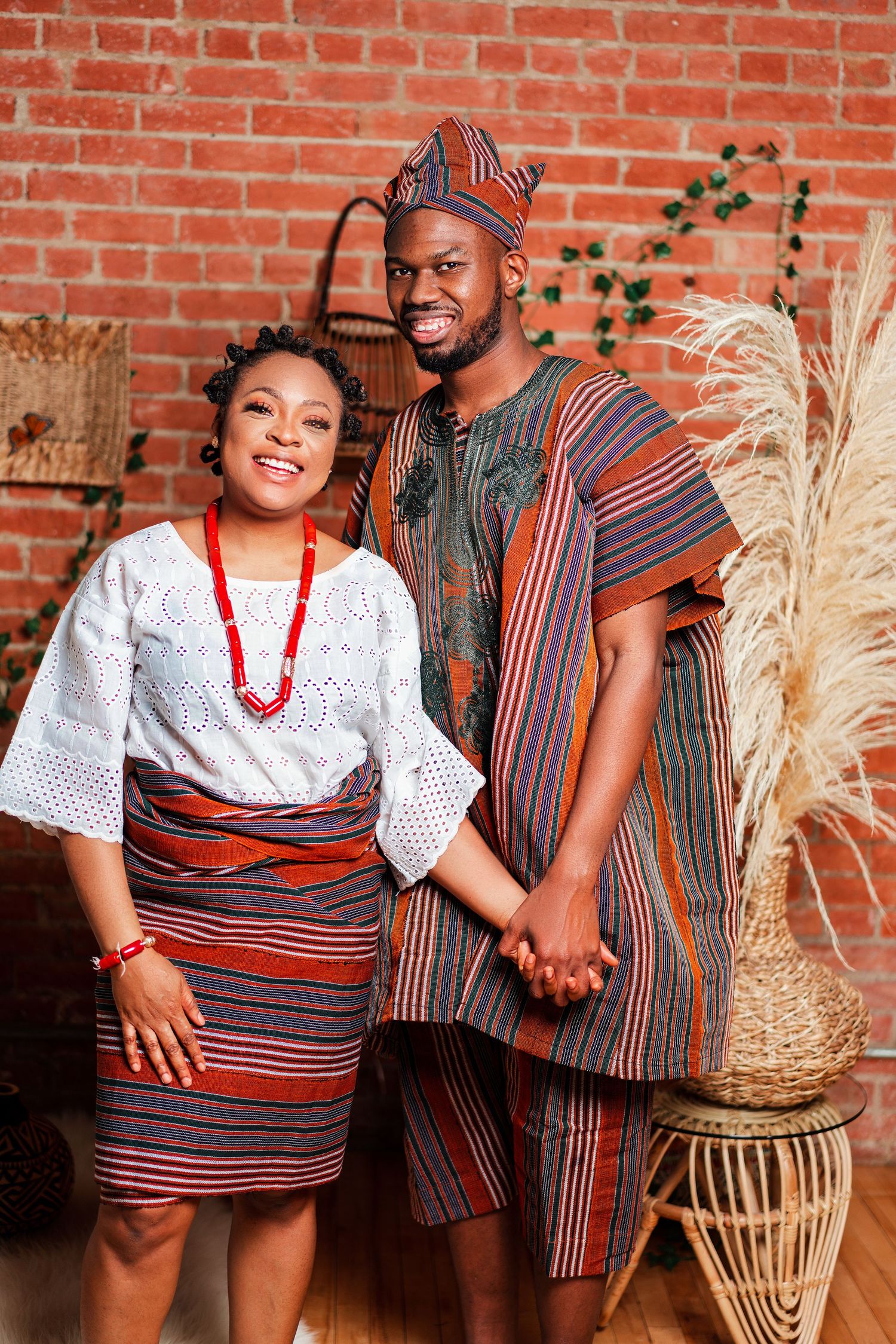 A couple in coordinated African-inspired attire poses together against a rustic brick wall setting.
