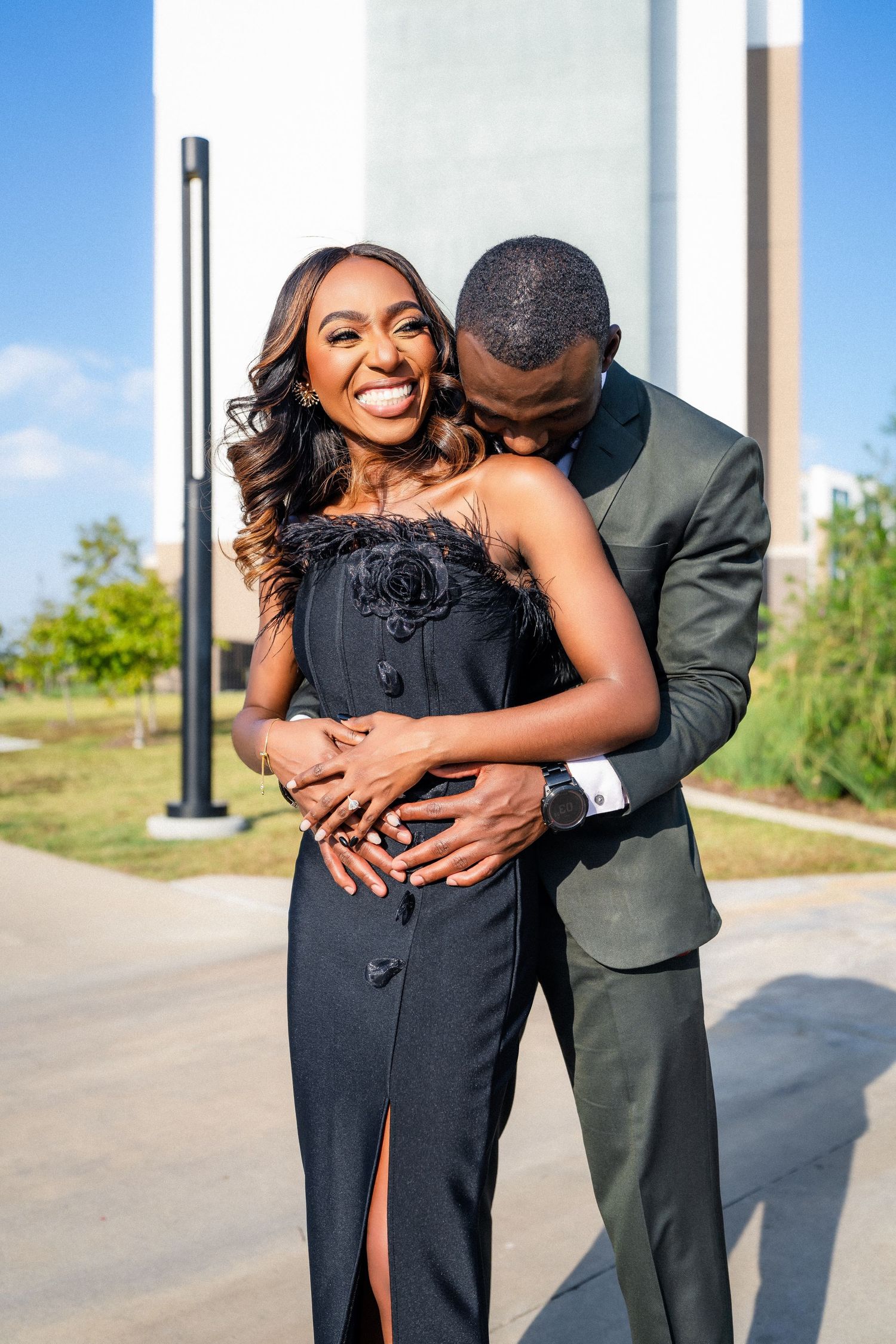 A couple in formal attire share playful embraces outdoors near architectural columns.