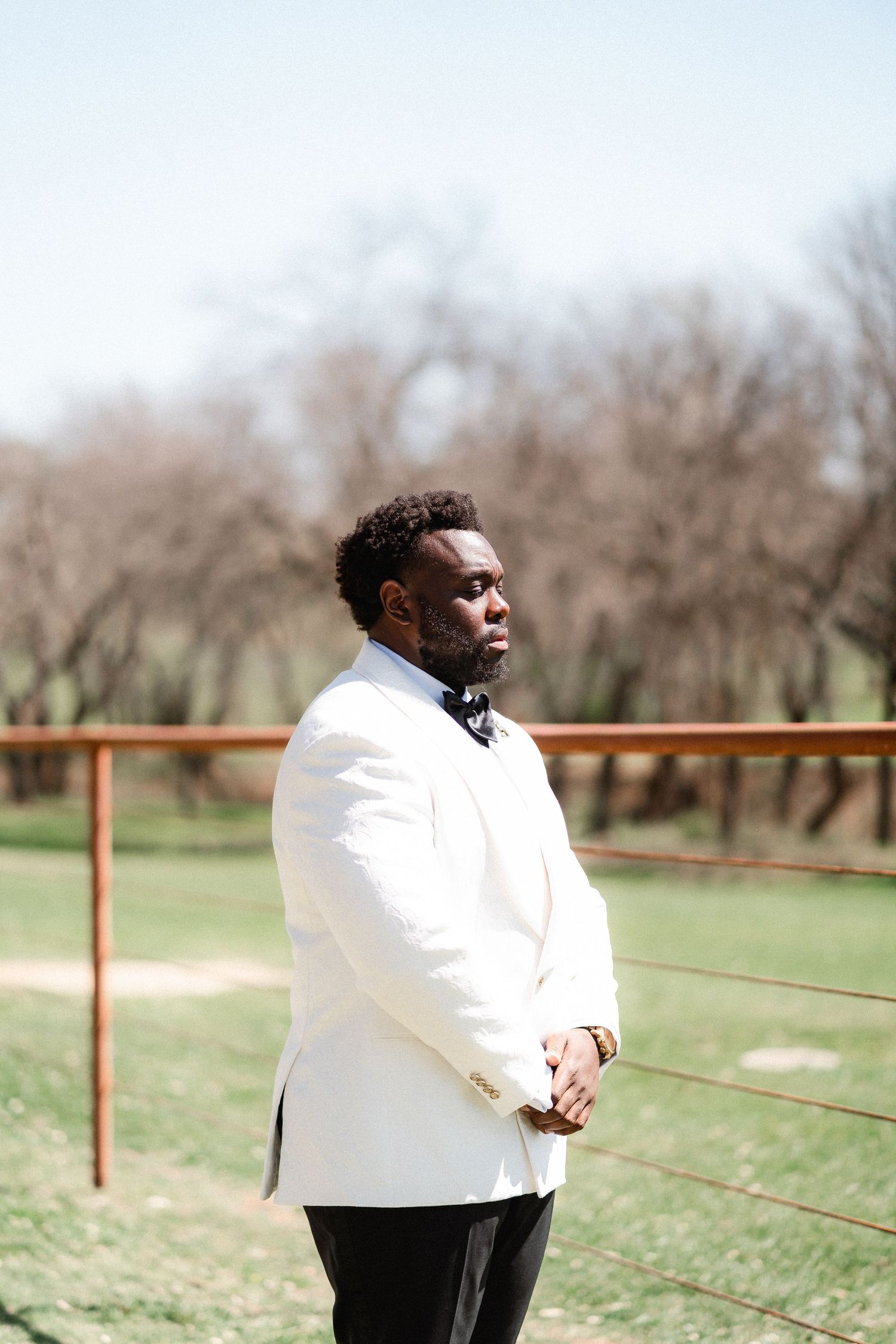 Groom in white jacket poses thoughtfully against rustic outdoor fence backdrop.