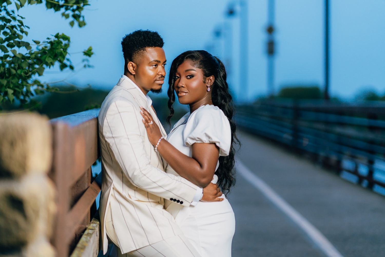 A romantic couple poses for engagement photos on a bridge during sunset wearing coordinated white outfits.