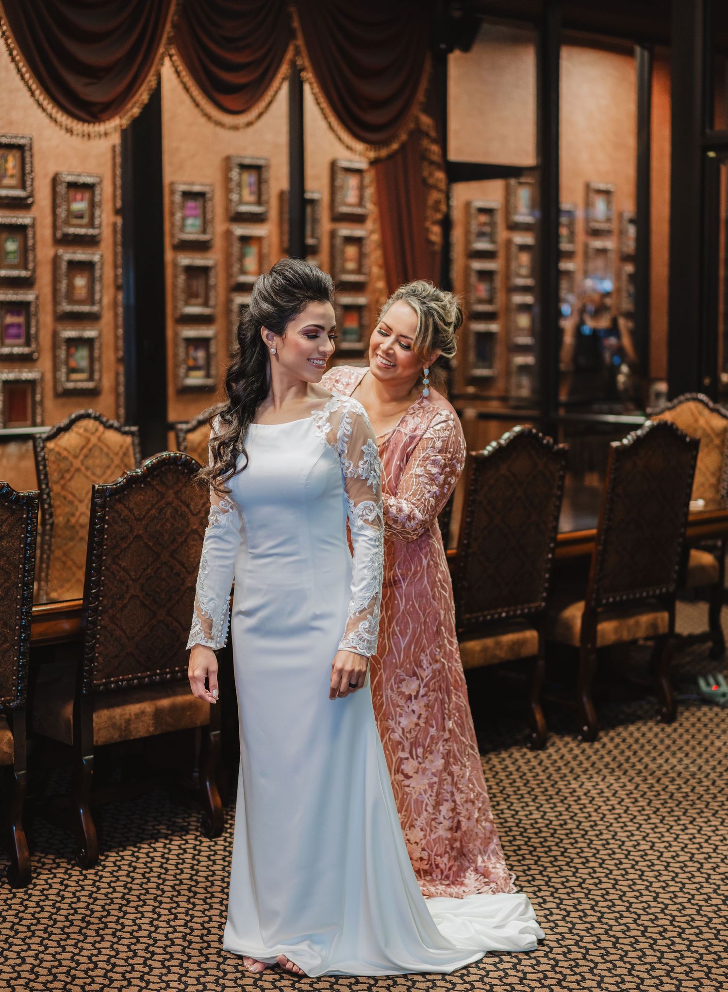 Two women in formal dresses pose together in an elegant room with ornate wall decor and velvet chairs.