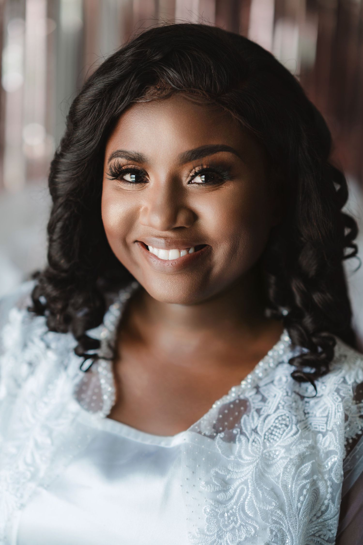 A radiant bride wearing a white lace dress smiles warmly in a portrait with soft lighting and elegant makeup.