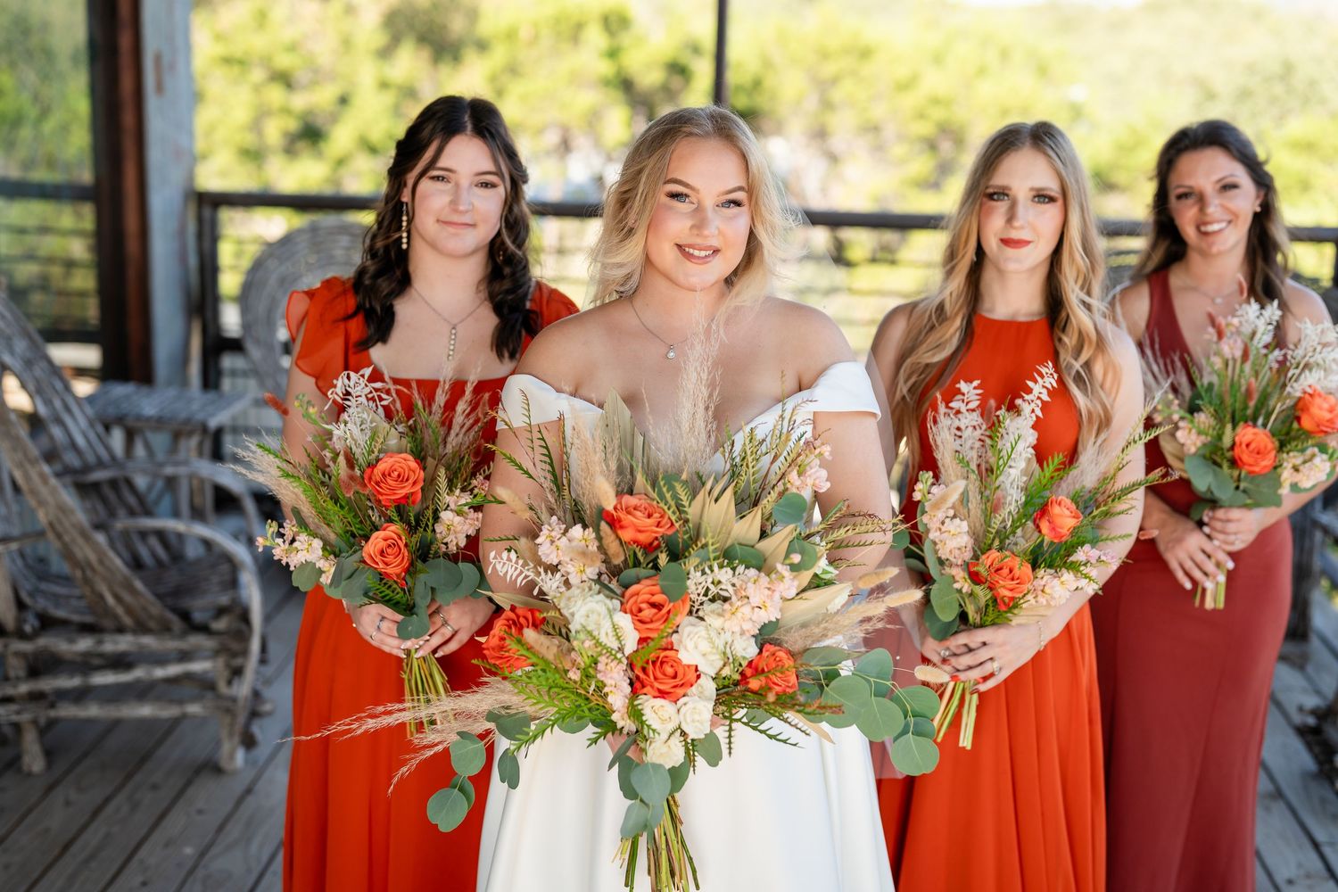 Wedding party in bright coral dresses holding matching floral bouquets on wooden deck.