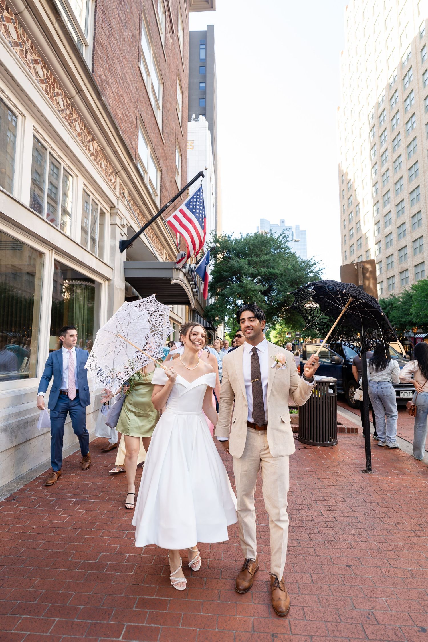 Wedding couple walks along brick sidewalk with American flags and umbrellas in background.