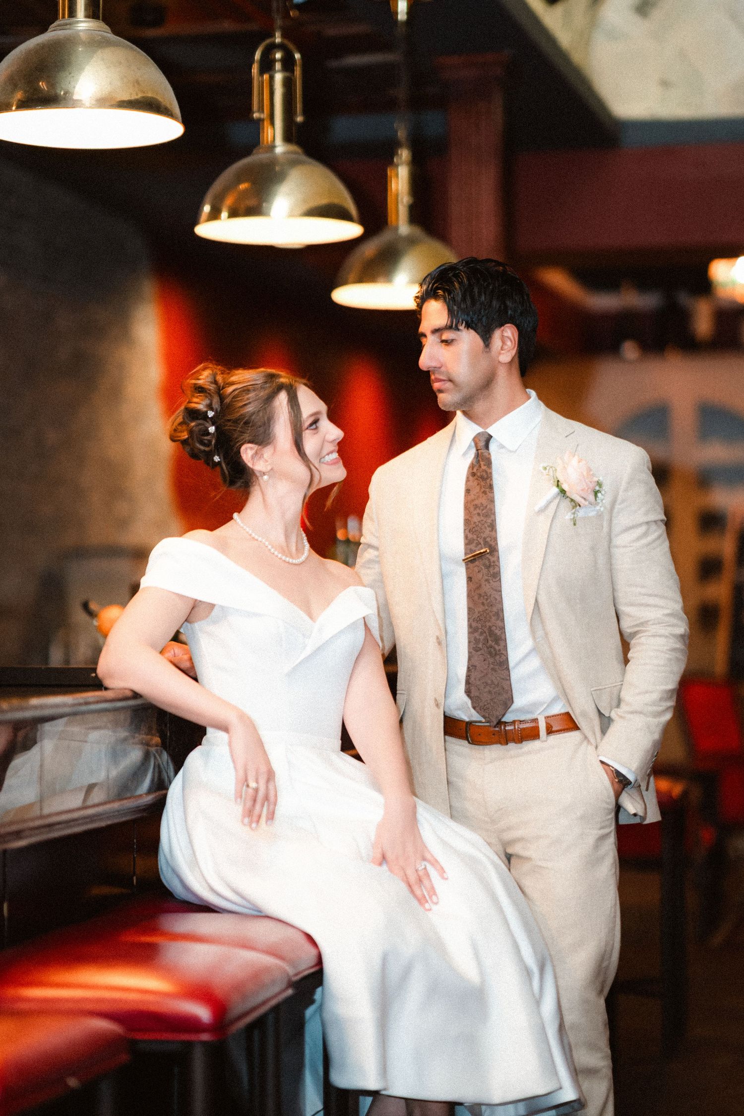 A couple in wedding attire poses together in an indoor venue with vintage lighting and red accents.