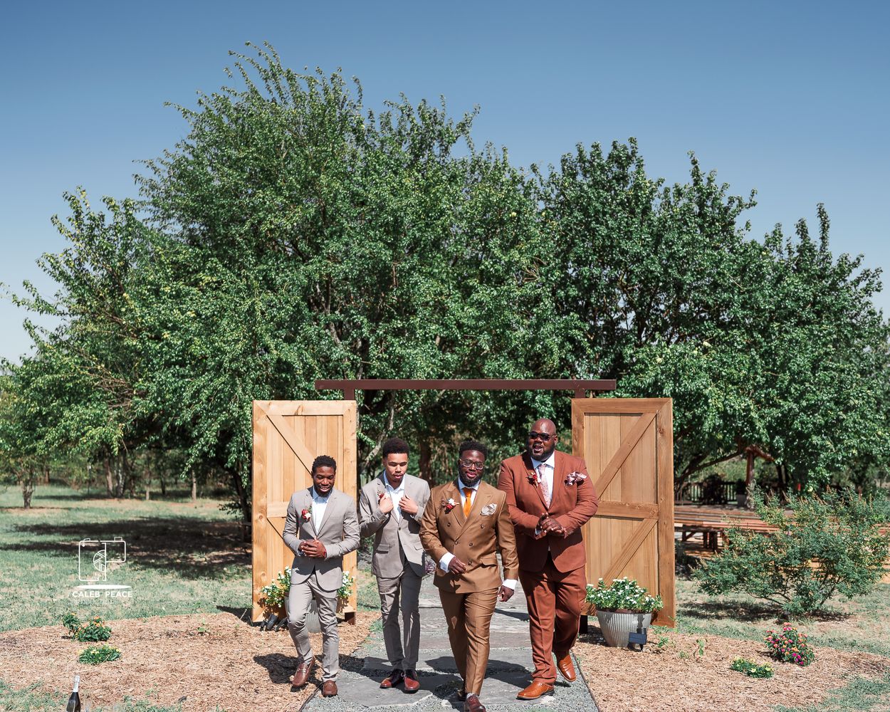 Wedding party poses for photos in front of rustic wooden doors in an orchard setting.