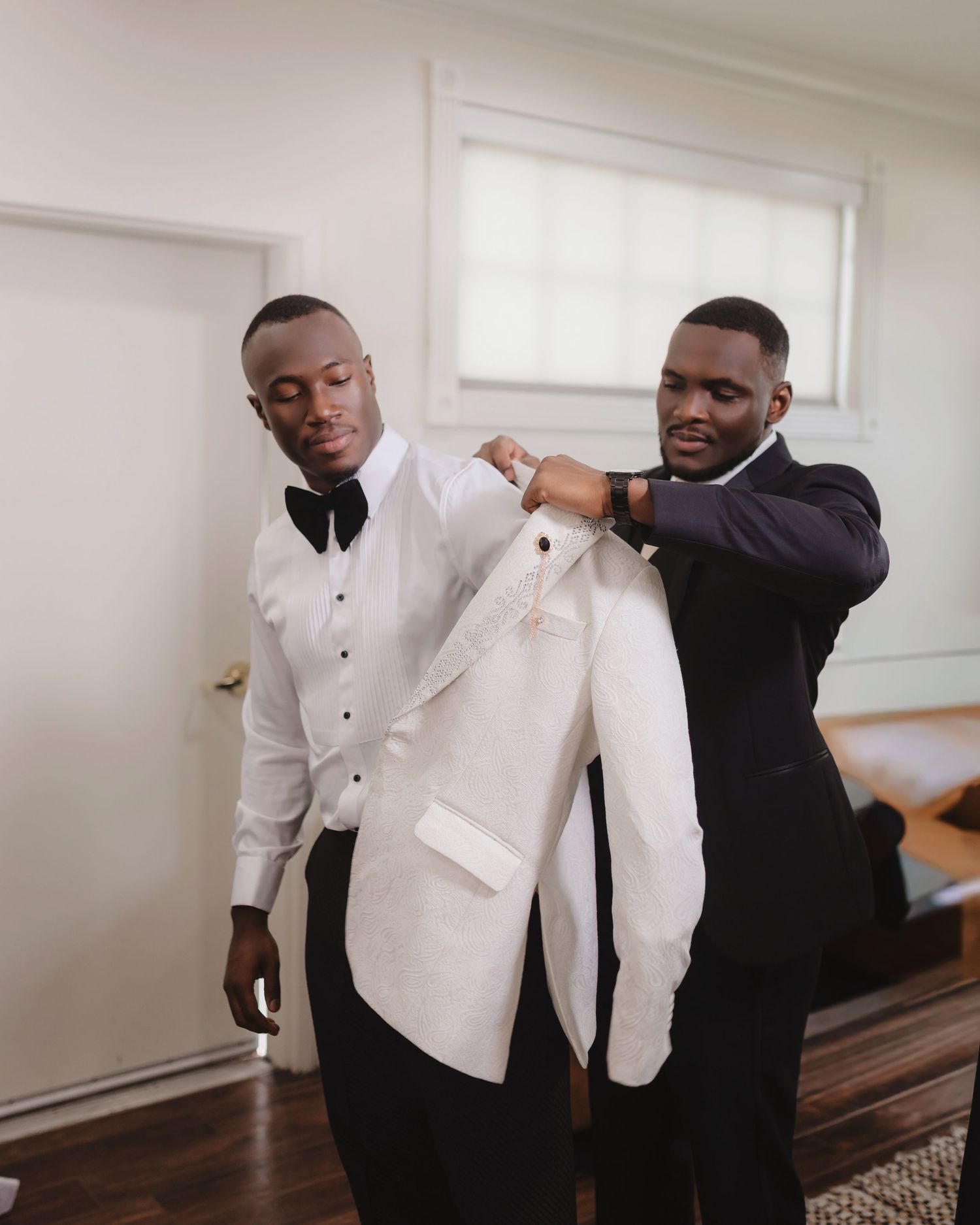 Groomsman helps adjust white tuxedo jacket during wedding preparation.