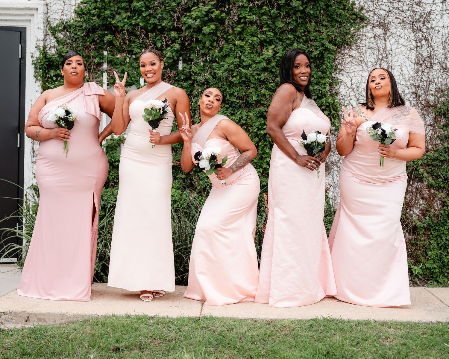 Bridesmaids in blush pink dresses pose together against a green ivy wall.