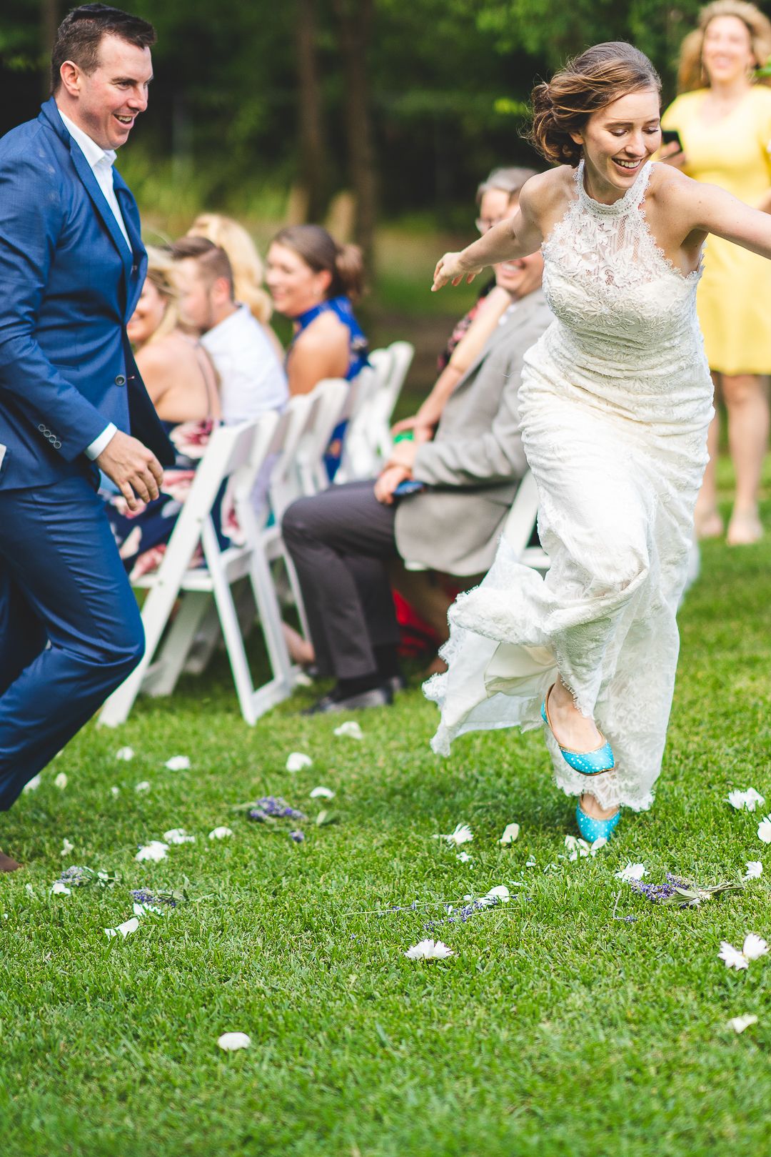 A newlywed couple runs joyfully down the aisle while guests seated in white chairs look on.