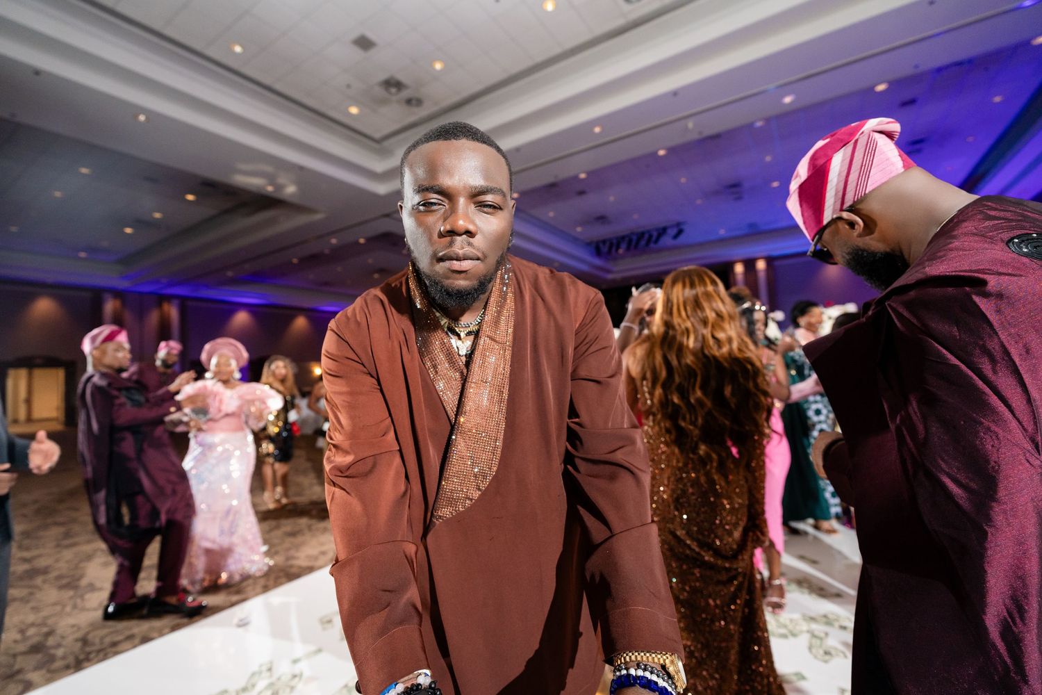 A guest in a brown suit stands at a wedding reception while other guests dance in the background.