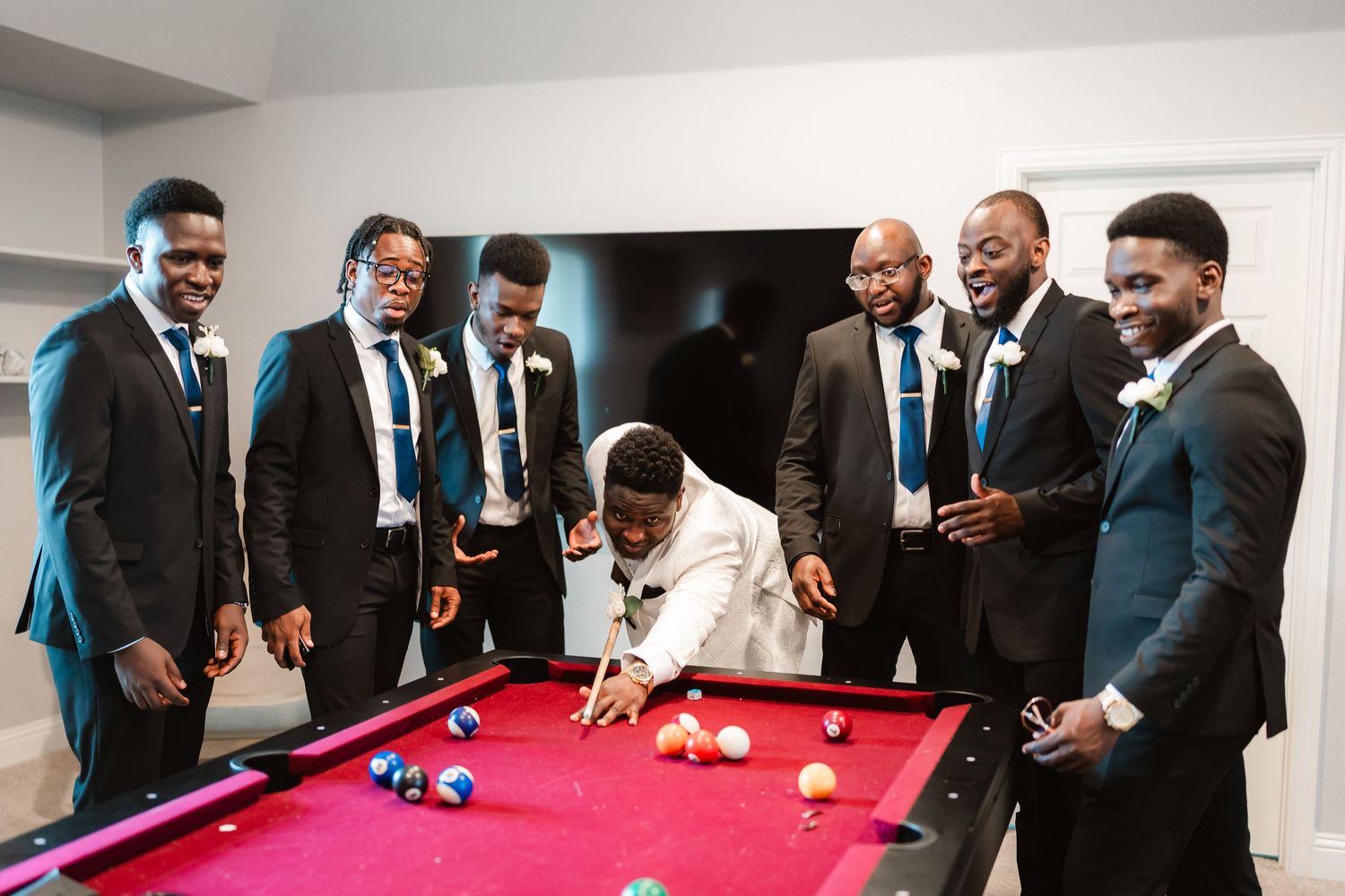 Wedding party guests in formal attire gathered around a red pool table enjoying recreational activities.