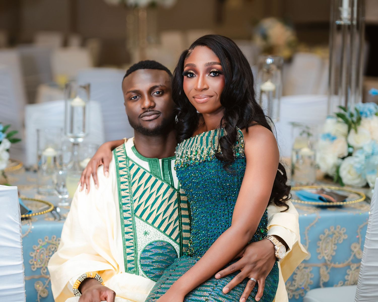 Couple in coordinating green and white formal attire pose together at elegant reception with table settings visible.