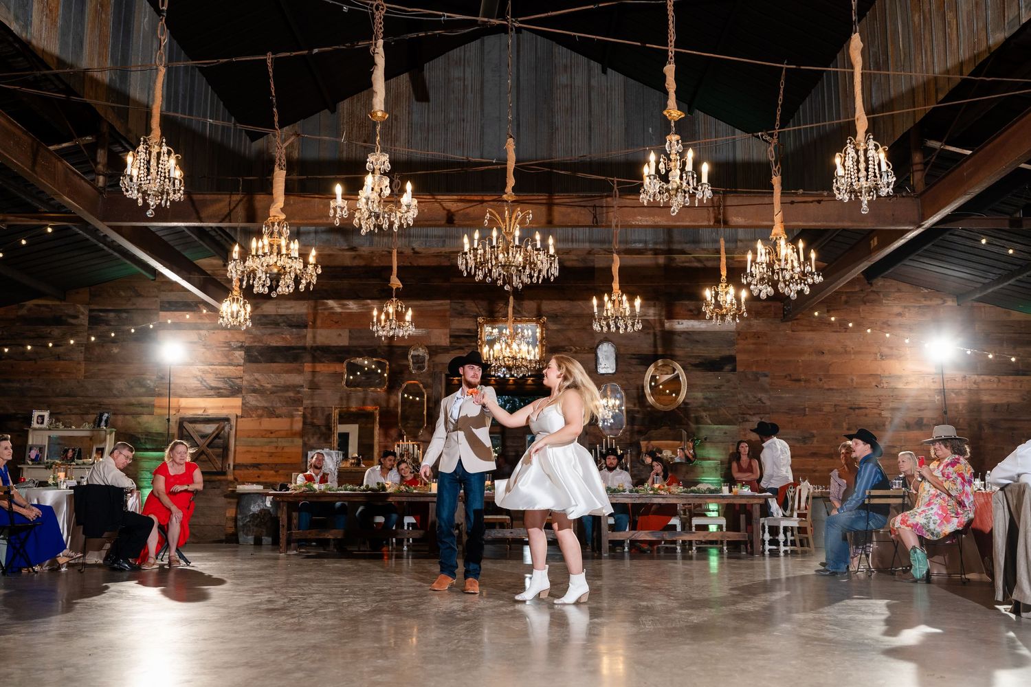 Panoramic view of a rustic barn wedding venue with multiple crystal chandeliers hanging from wooden beams.