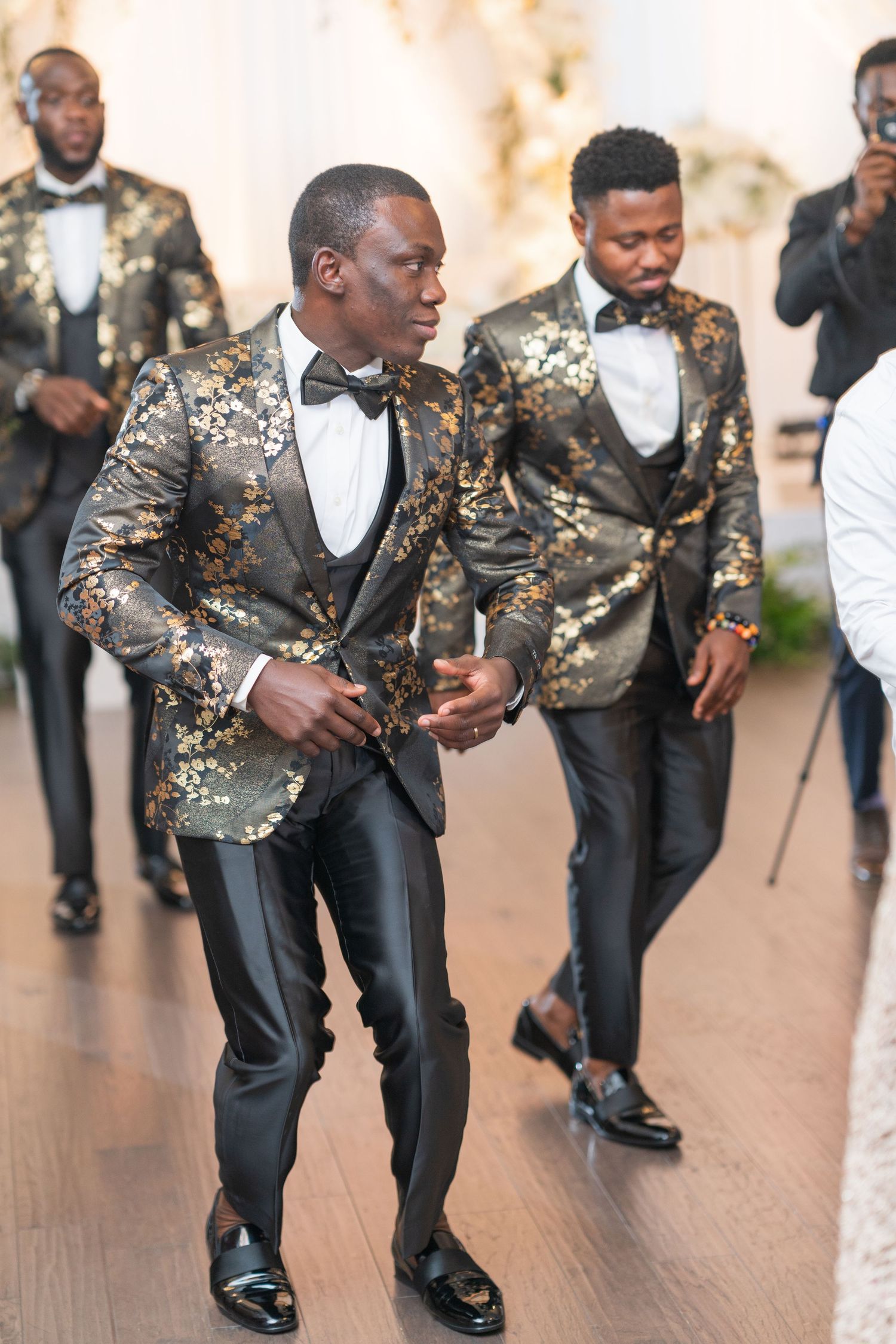 Groomsmen in stylish gold and black floral tuxedos dance and celebrate at a wedding reception.
